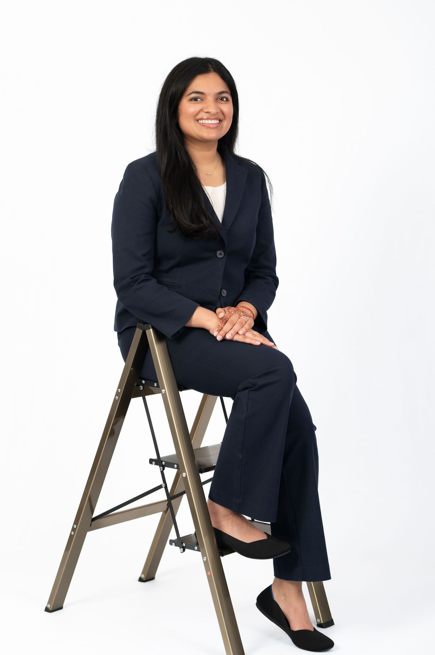 full length business portrait of a young woman in a navy business suit and white blouse, posing seated on a metal ladder on a white backdrop