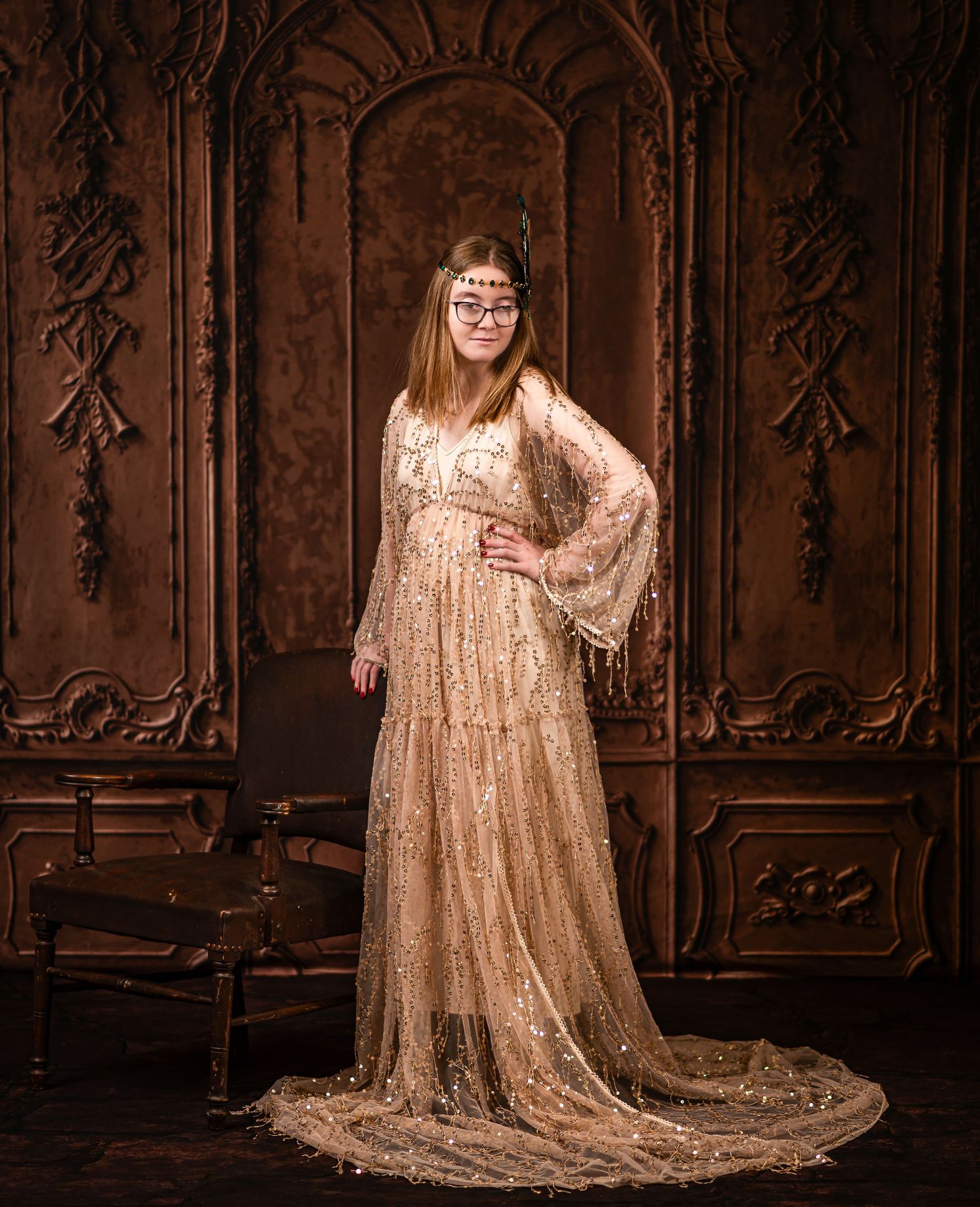 classical style studio portrait of a young woman in a floor length gold sequin gown posing in front of an ornate carved paneled wall