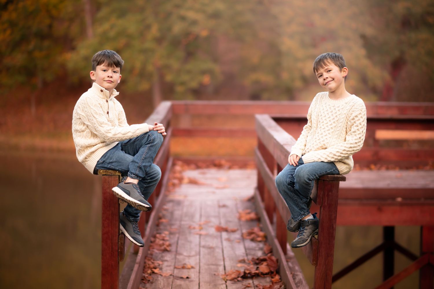 two boys in cream sweaters and blue jeans pose for a portrait on opposite sides of a wooden fishing pier will fall leaves in the background