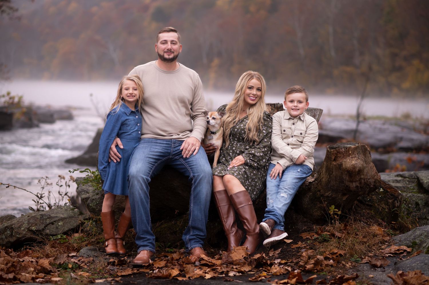 a family of four in fall colors poses on a large fallen log in front of the river with fog and trees in the background; shot in Valley Falls