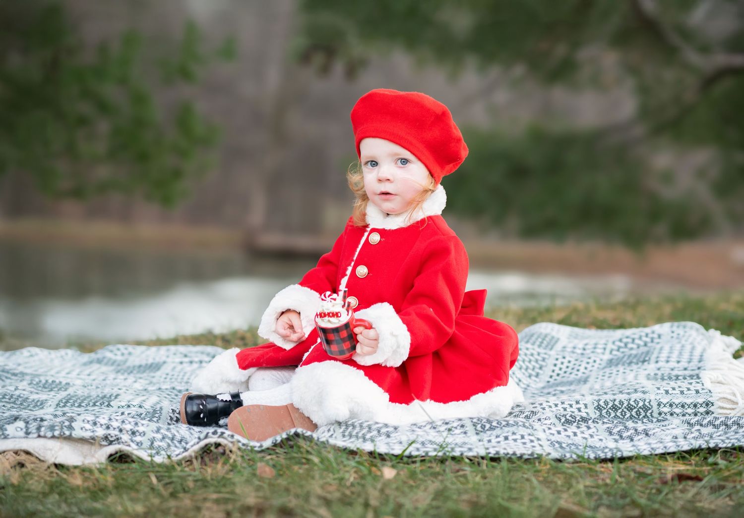 Christmas portrait of a toddler in a fancy red and white fur coat and little black dress shoes, seated on a blanket outside with evergreens