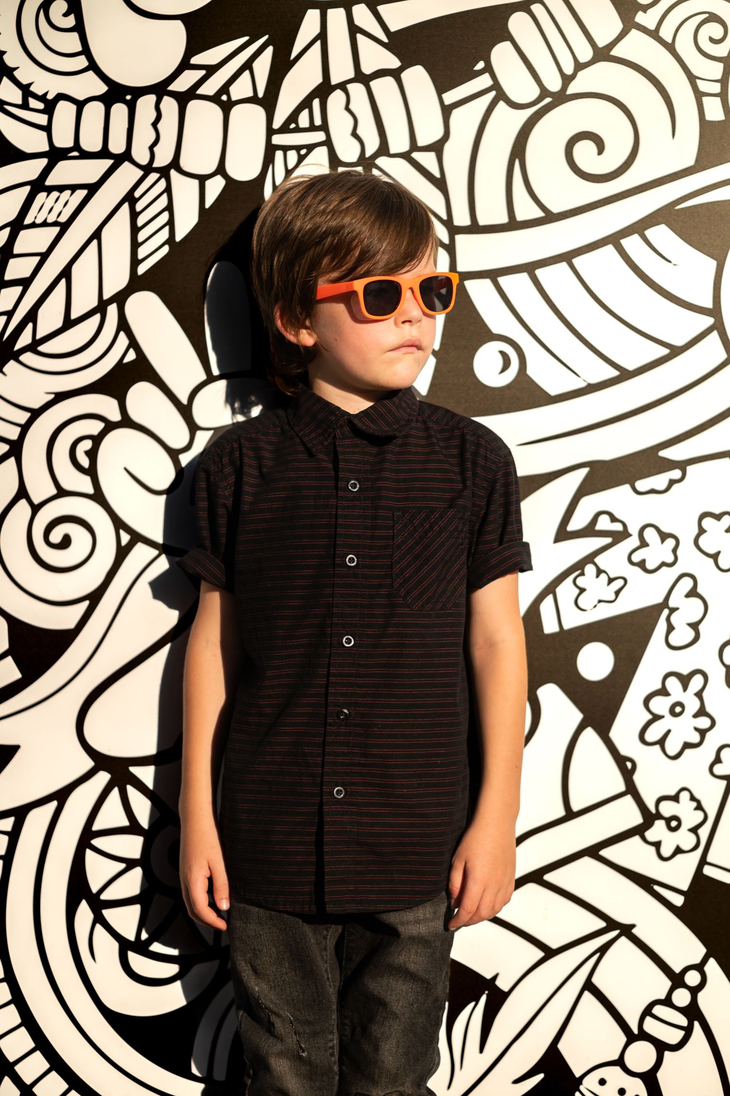 a young boy in all black with orange sunglasses lounges against a black and white modern art mural and looks away from the camera
