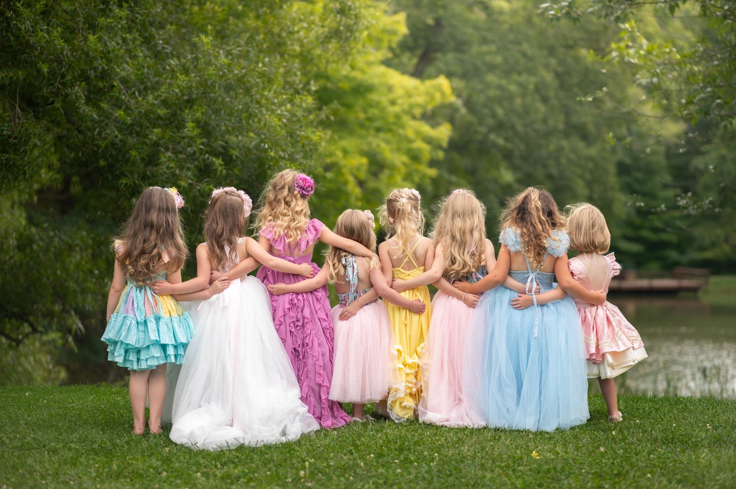 a group of little girls in fancy fluffy dresses stands together with arms around each other looking out over a beautiful pond