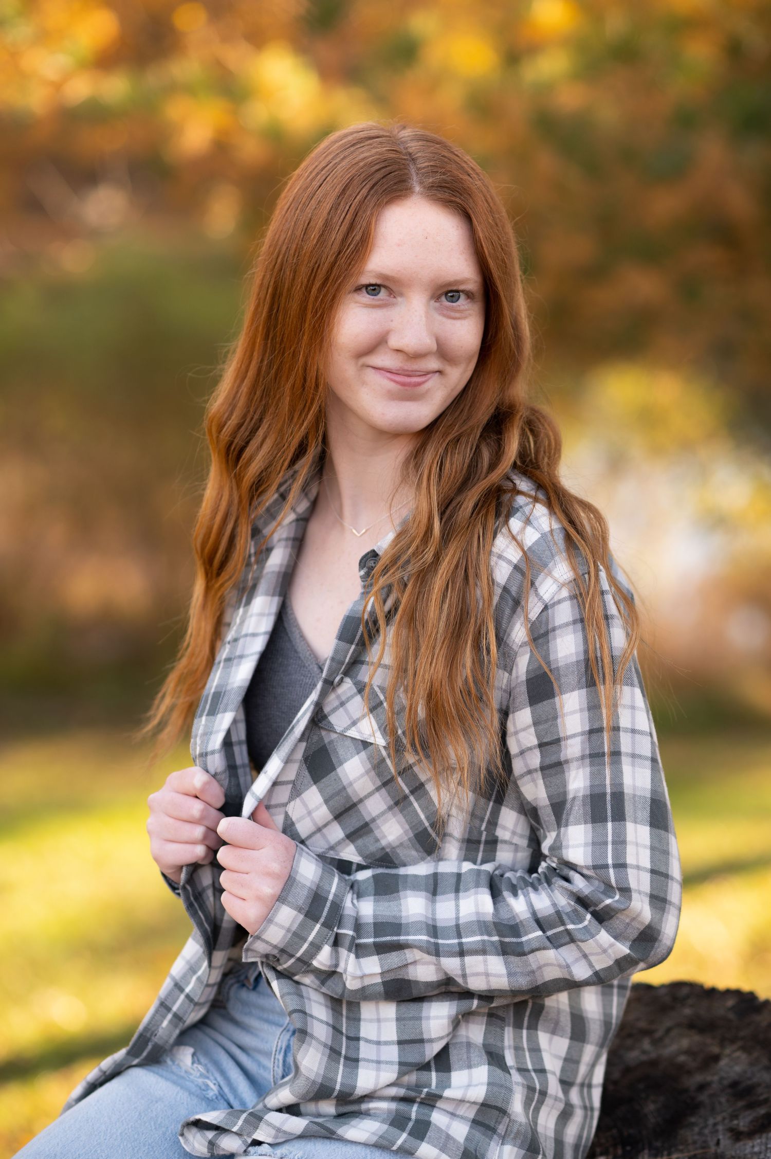 redhaired senior student in a plaid shirt poses and smiles on a stump with sunlit trees, grass, and a pond in the background