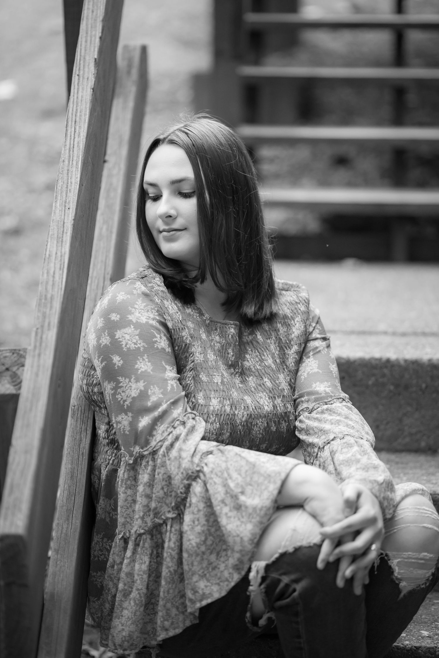 black and white portrait of a young woman with dark hair in a boho top, seated on a set of old wooden stairs and looking away from the camer