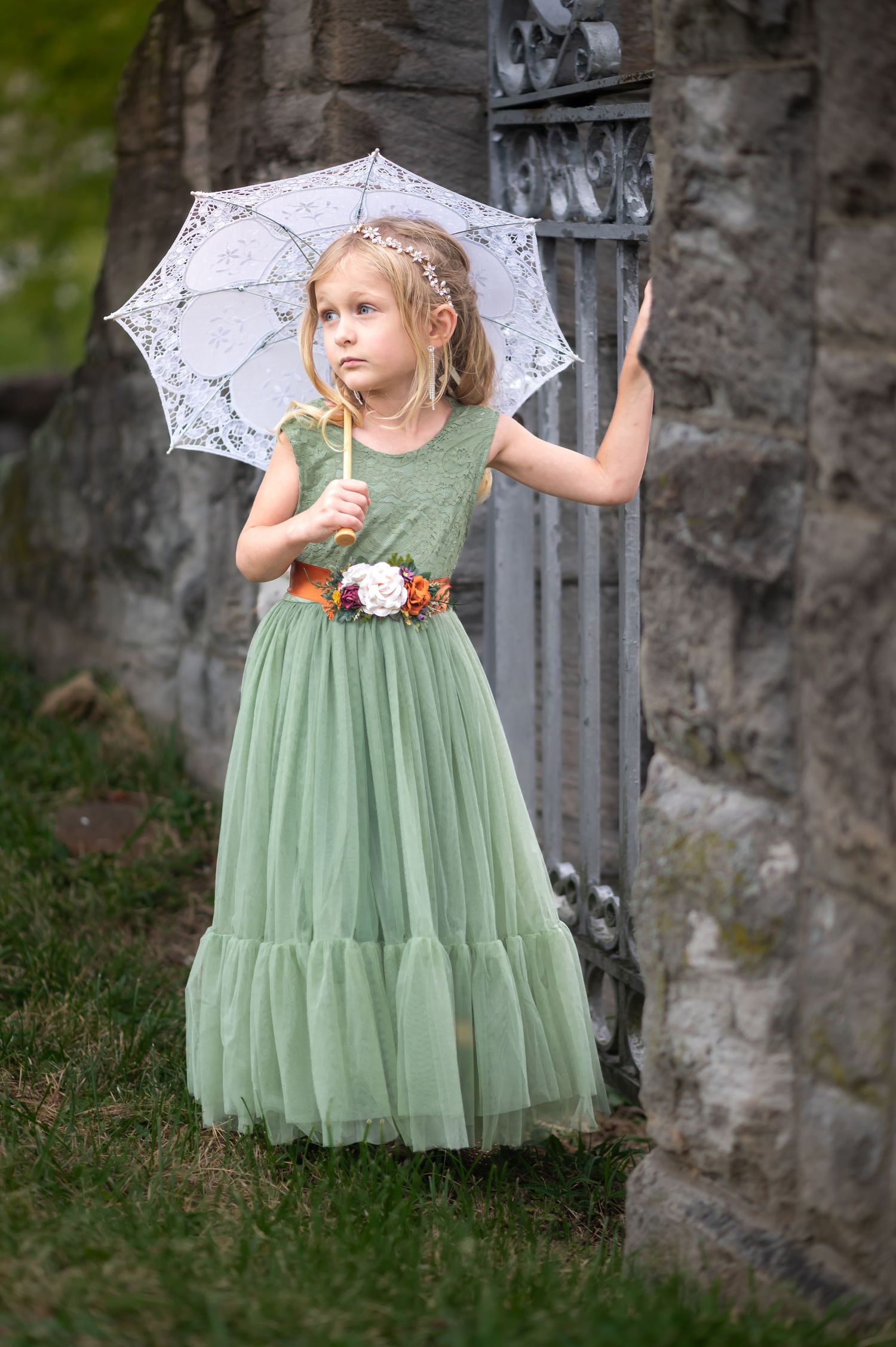 a little girl in a fluffy floor length green dress with a white parasol poses in front of an old antique iron gate and stone arch