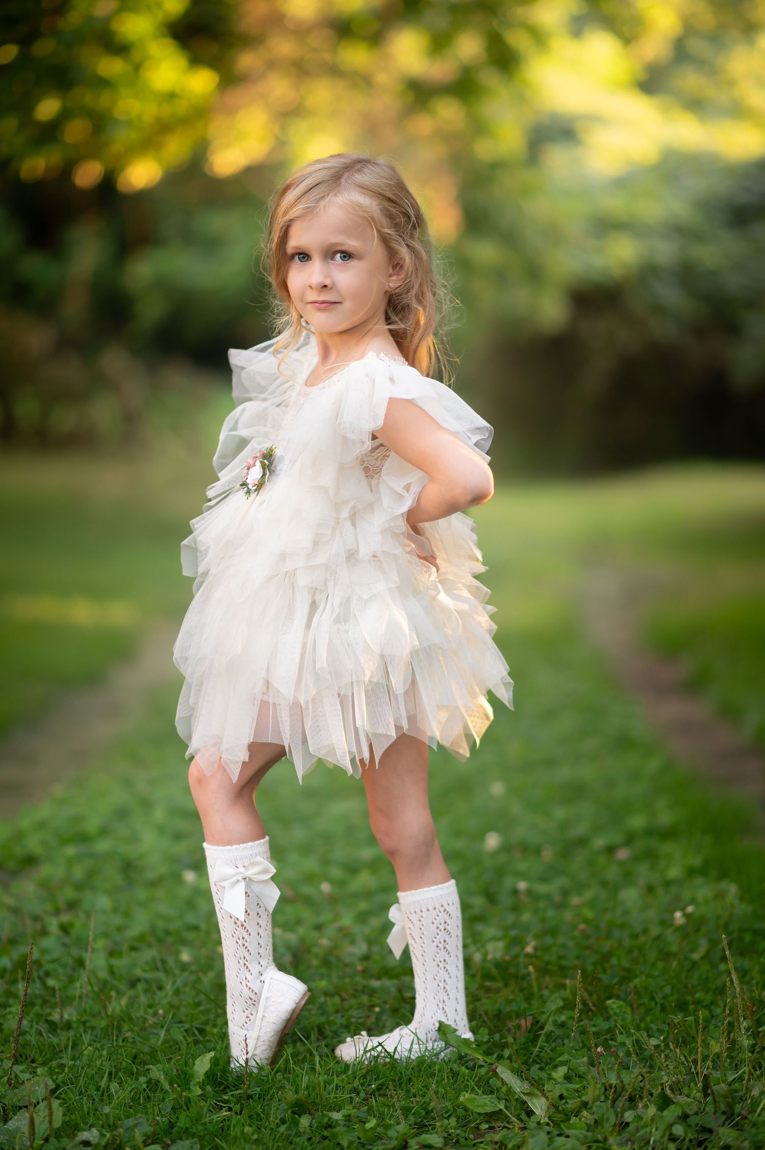 a pretty little blonde girl in a fluffy frilly white lace dress and white stockings poses with one hand on her hip.