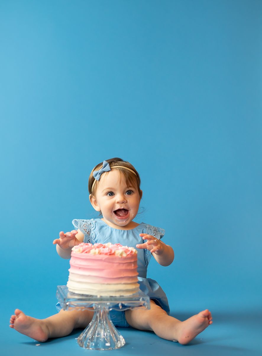 a one year old girl in a blue dress sits on a blue studio background with her big pink and white birthday cake on a crystal pedestal