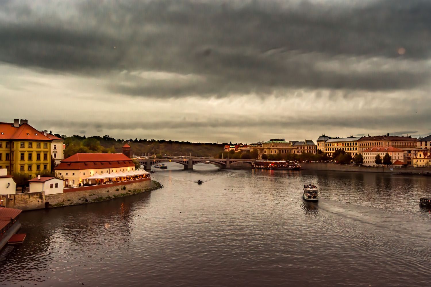 Stormy skies over Prague's Vltava River with historic buildings, bridges, and a boat navigating the dark waters.