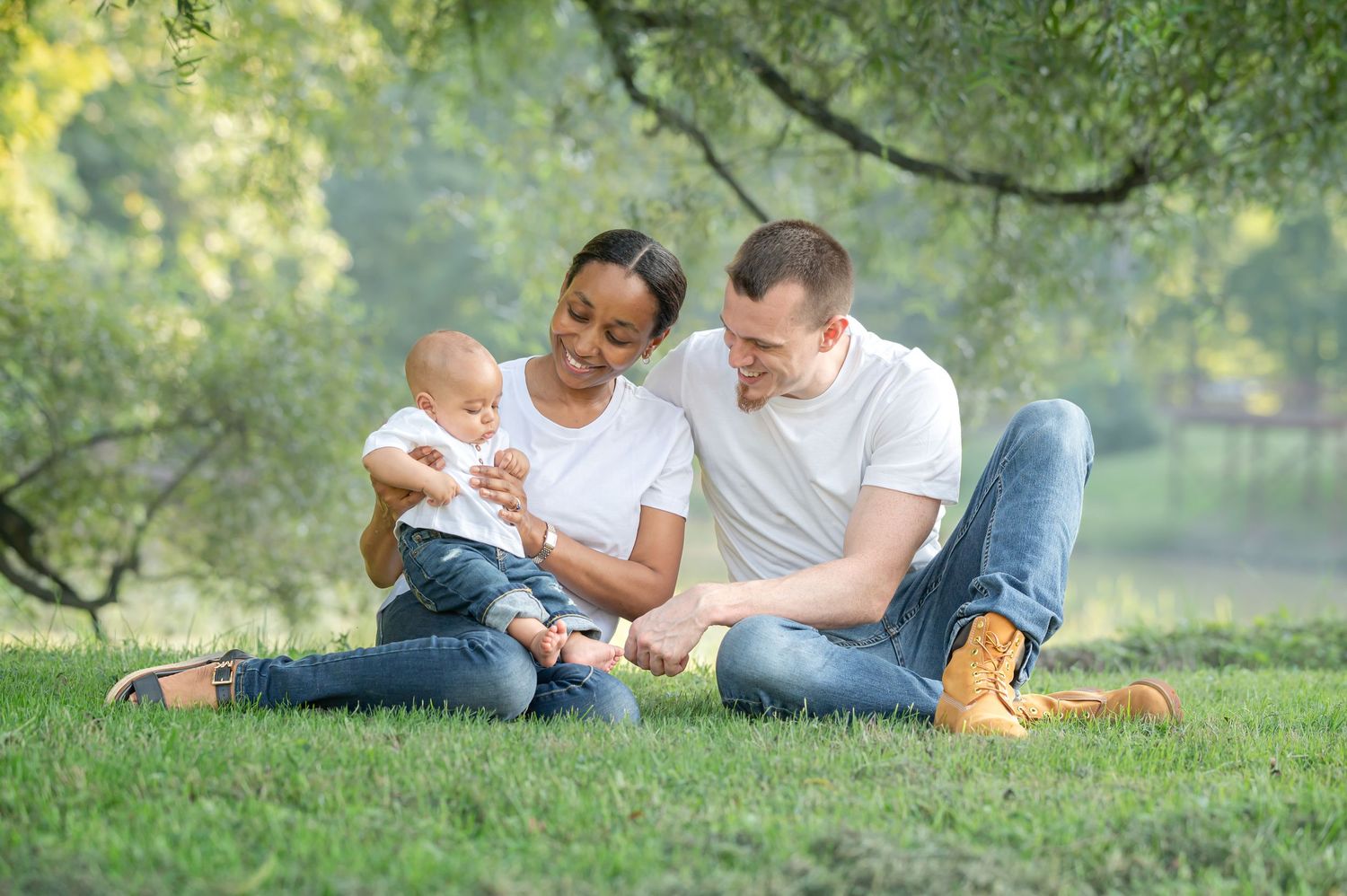 a young couple sit in the grass with their baby boy and smile at him, with soft green and gold trees in the background