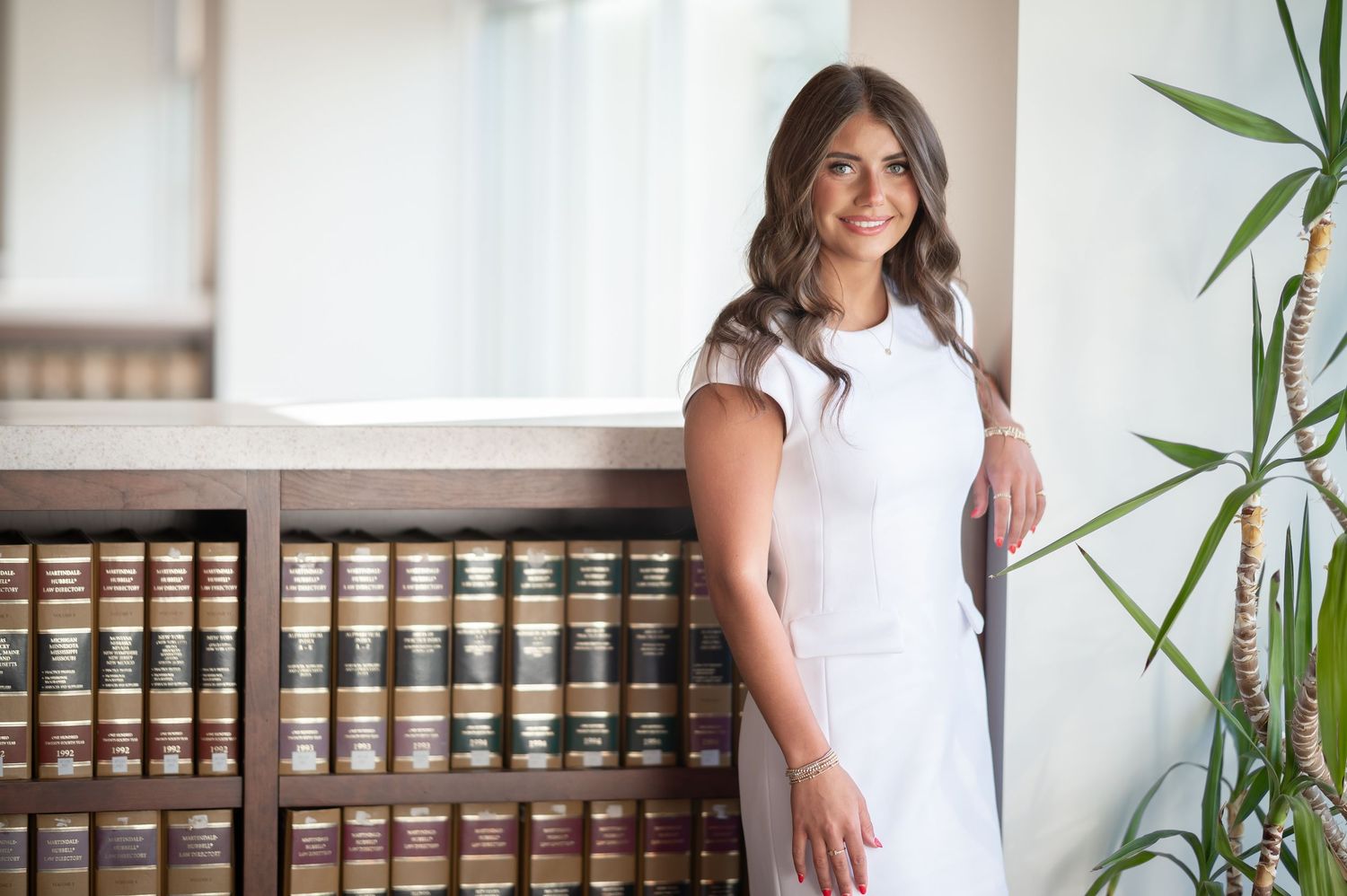 a WVU school of law graduate student in a white dress poses for a portrait beside a long shelf of law books in the library