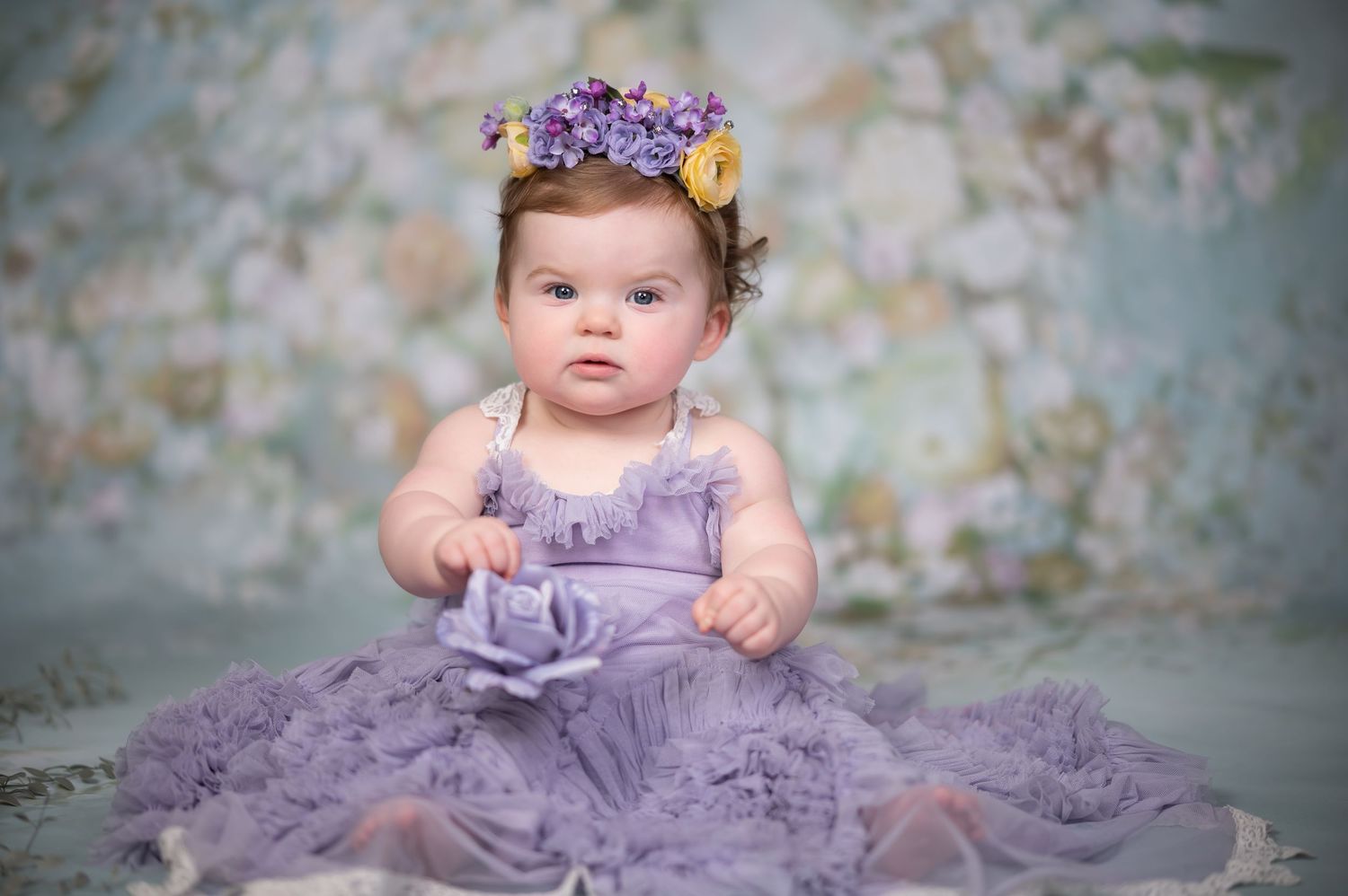 a toddler girl in a frilly lilac dress and purple and yellow floral crown sits on a green and purple floral background for a studio portrait