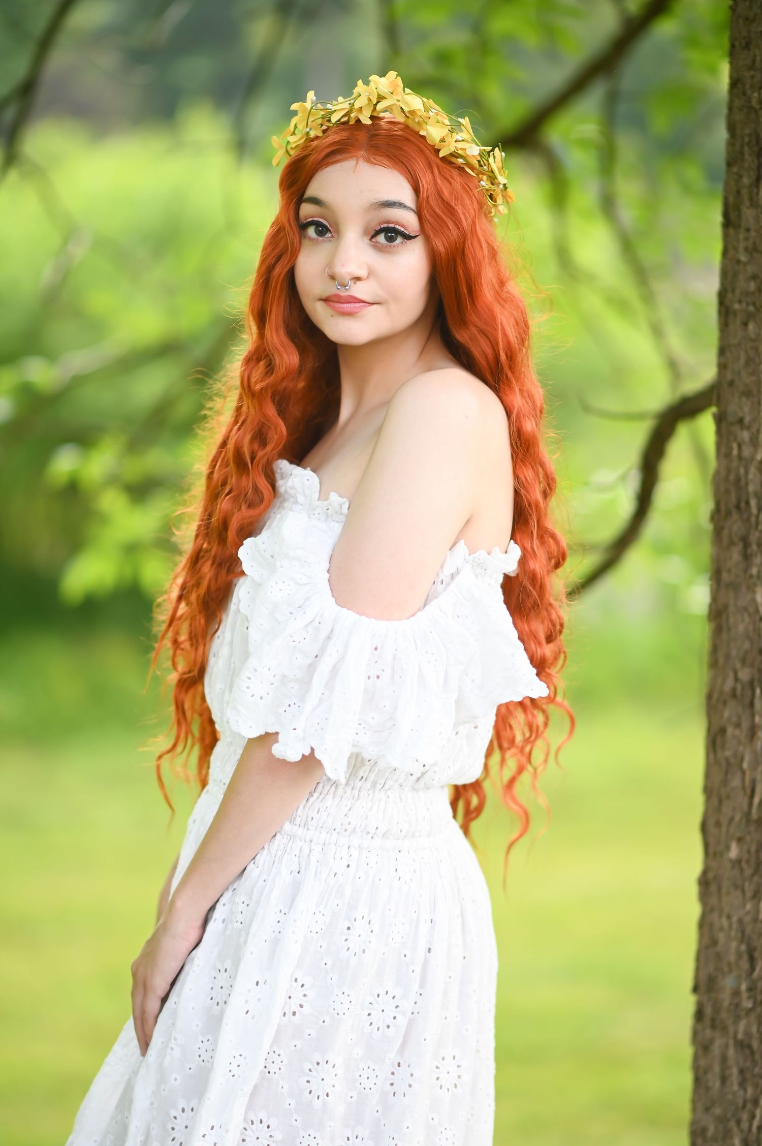 a young woman with long red hair and a white dress poses under a large tree, with lush green grass in the background