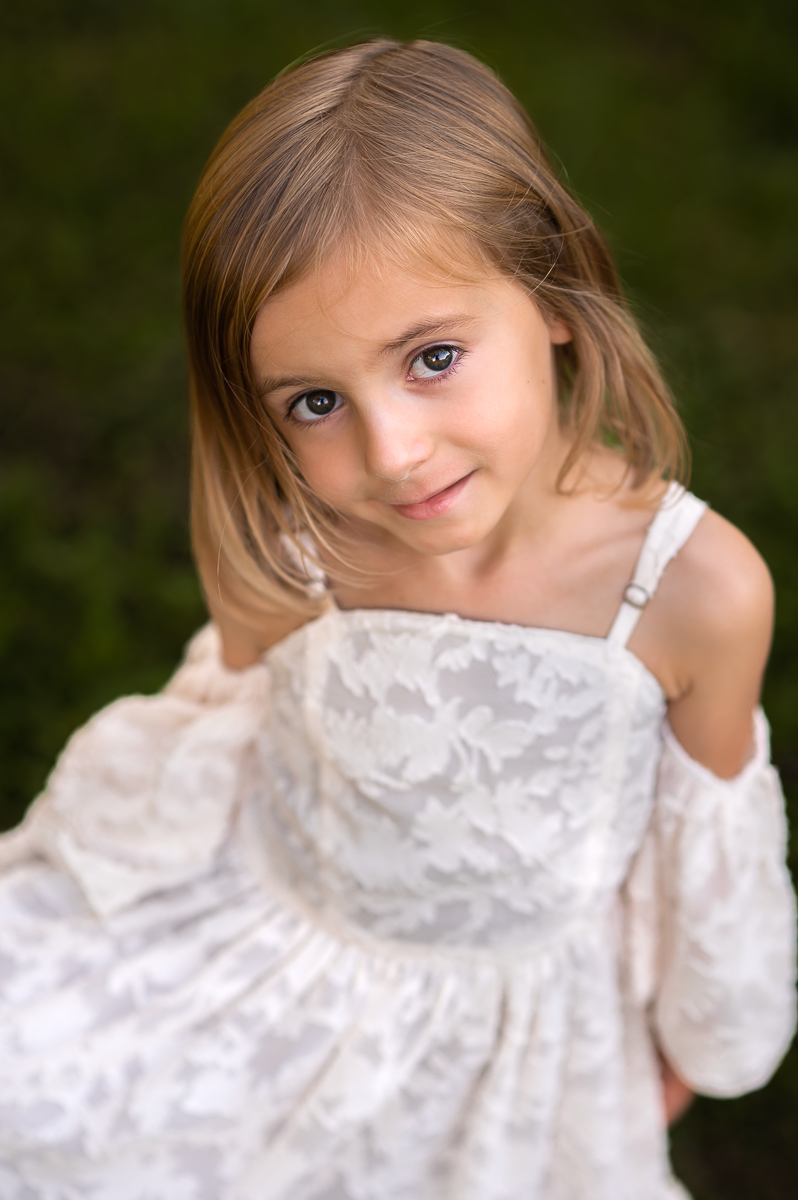 a little girl with brown hair and brown eyes in a white dress sits in the grass and smiles shyly up at the camera for a portrait