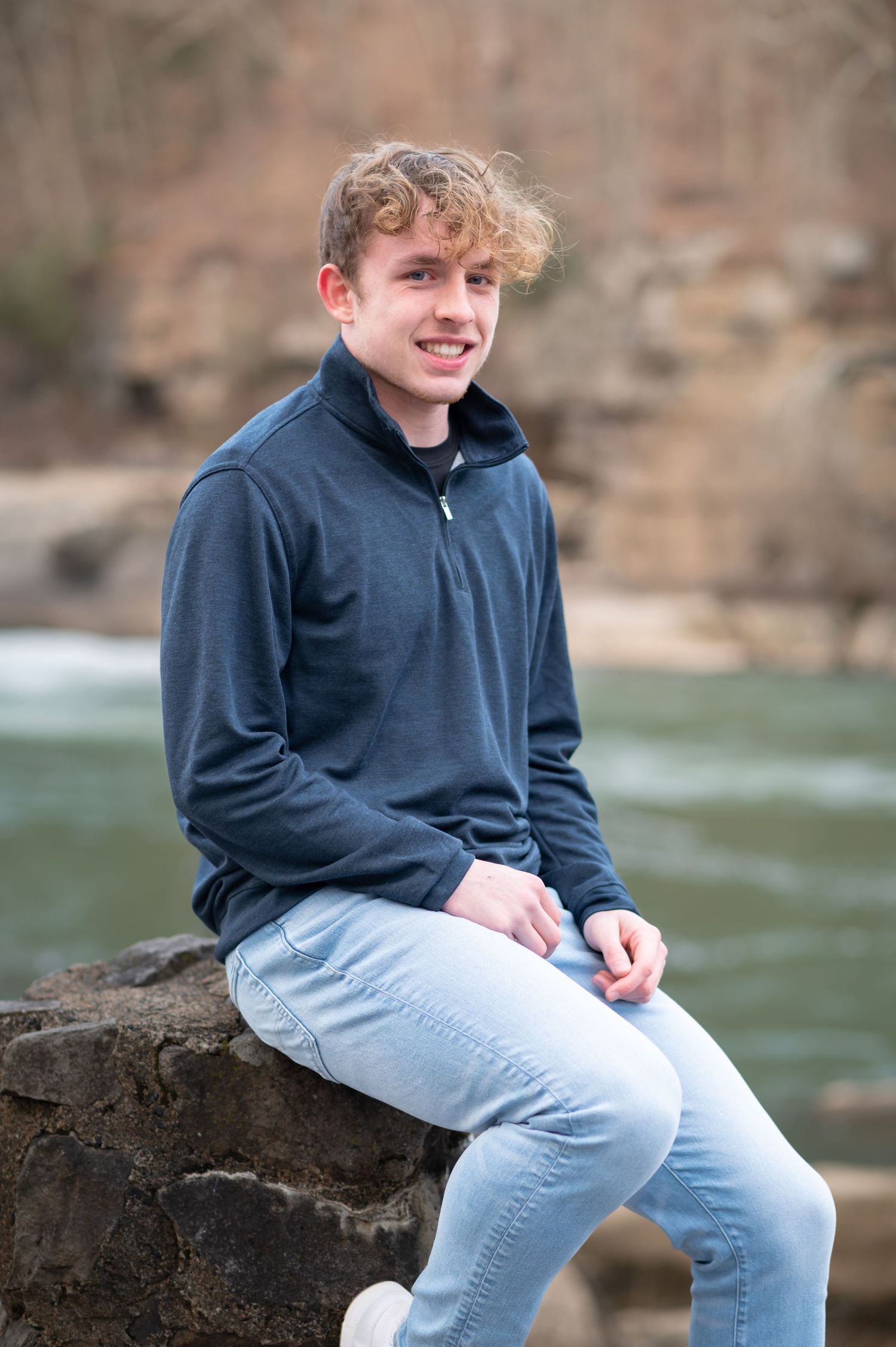 a young man with curly hair and a blue jacket sits on a large rock in front of a river, with waterfalls and trees in the background