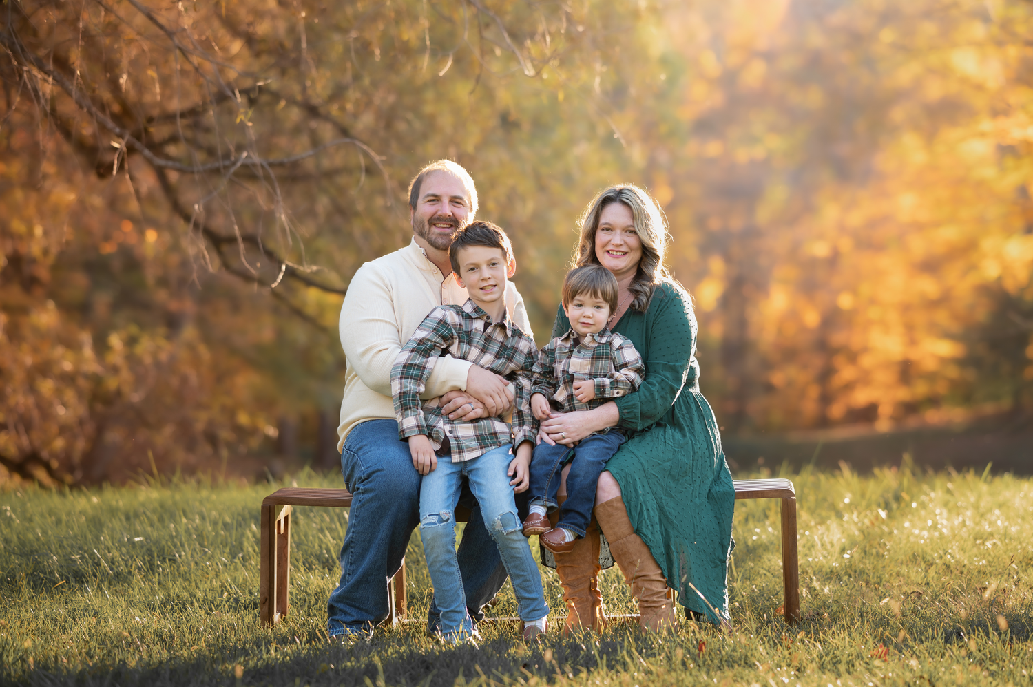 a family of four in green and cream tops and blue jeans poses for a family portrait on a bench with brilliant gold foliage in the background