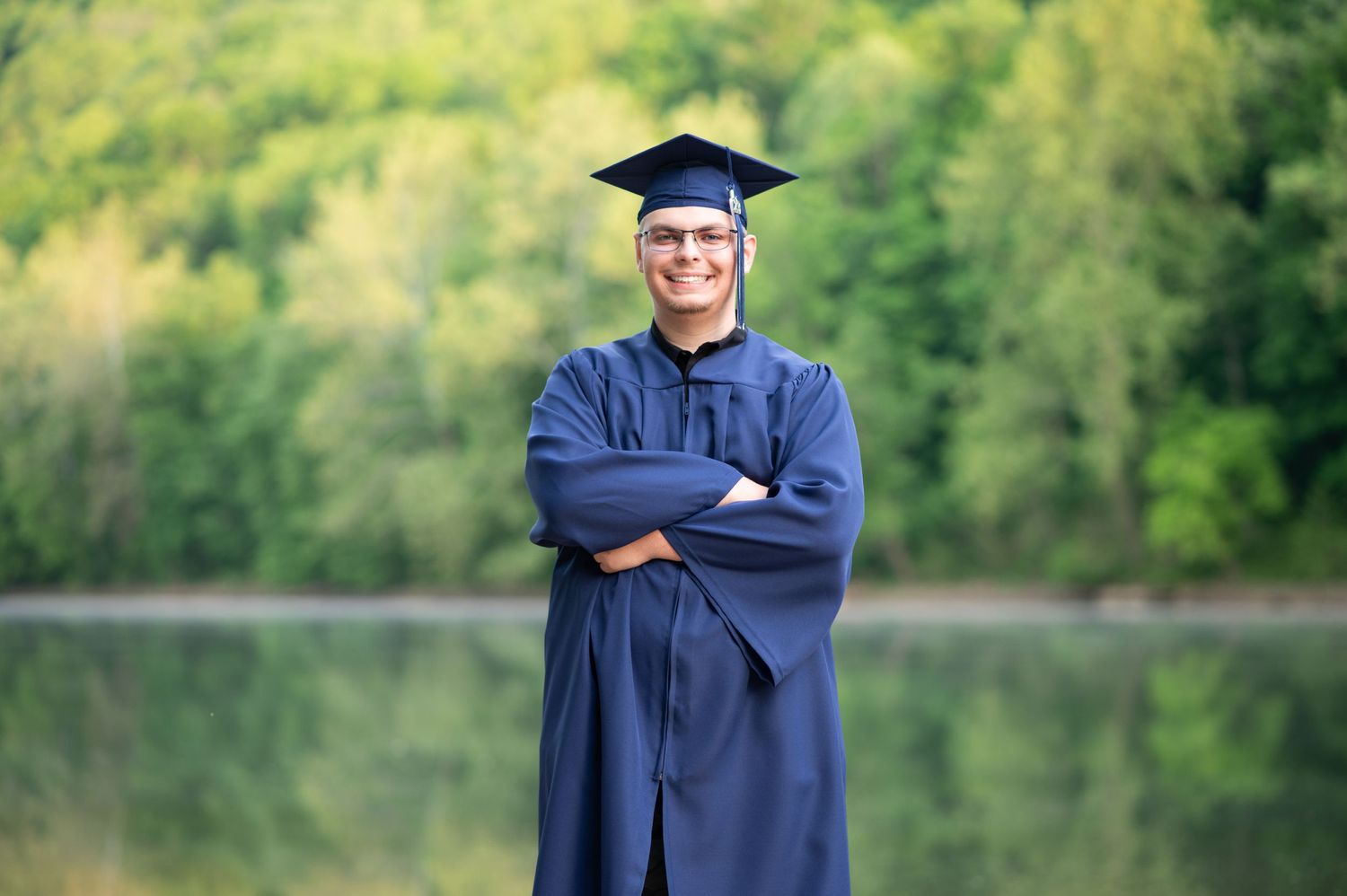 Young high school graduate in cap & gown poses for a senior portrait with arms crossed and a smile in front of the Tygart River in Fairmont