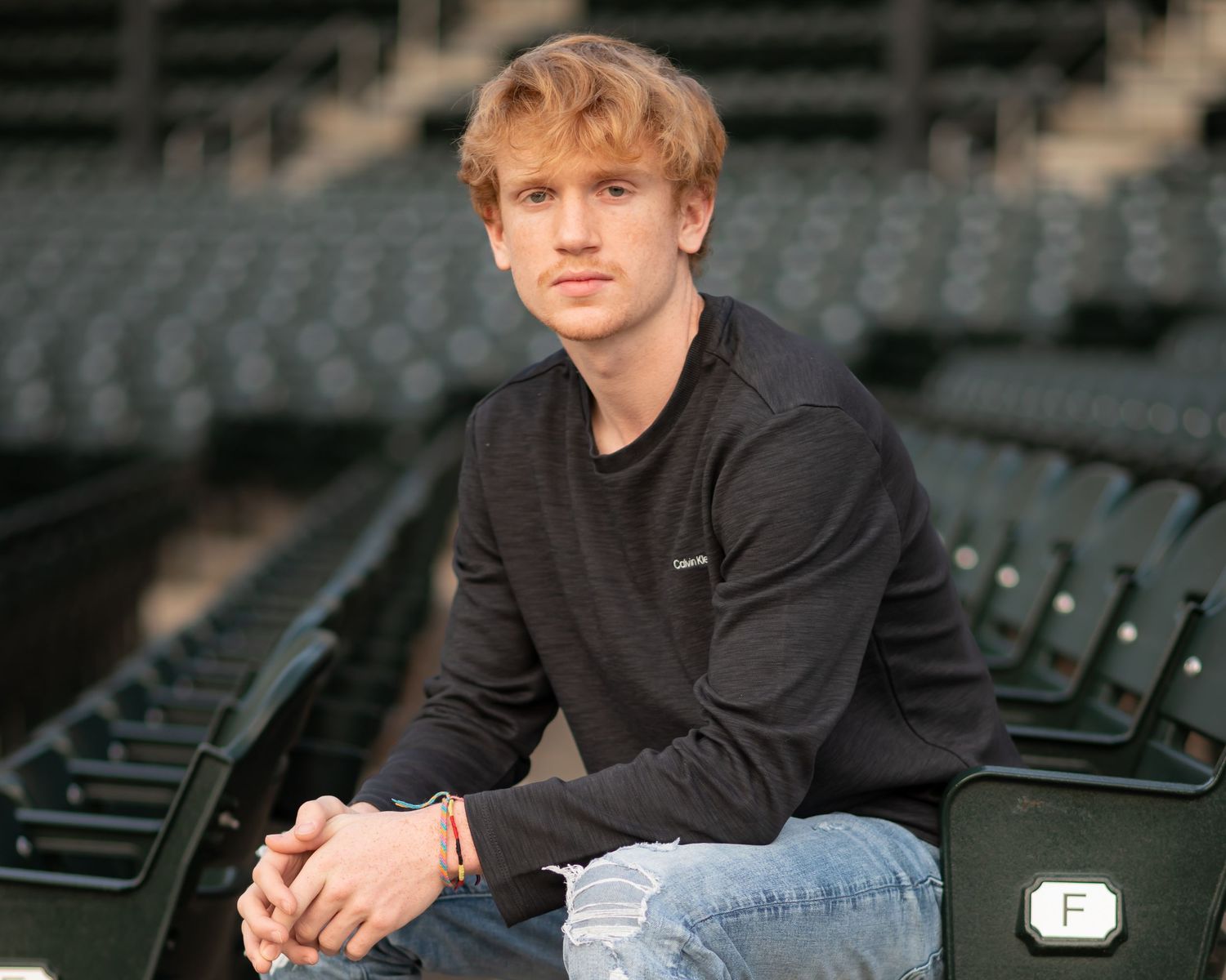 portrait of a young man in a black shirt and jeans, seated in empty stadium seating and looking into camera with a thoughtful expression