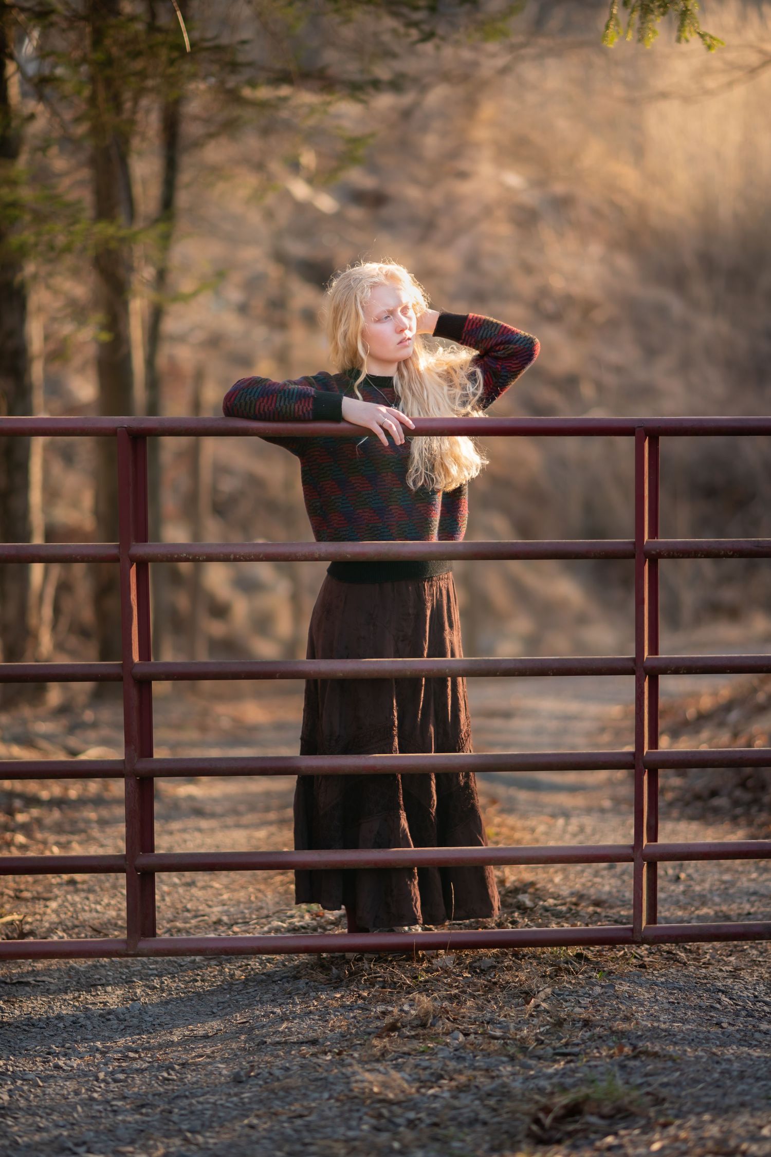 environmental senior portrait of a young blonde woman in a long black dress leaning against an old red gate with a country road behind her