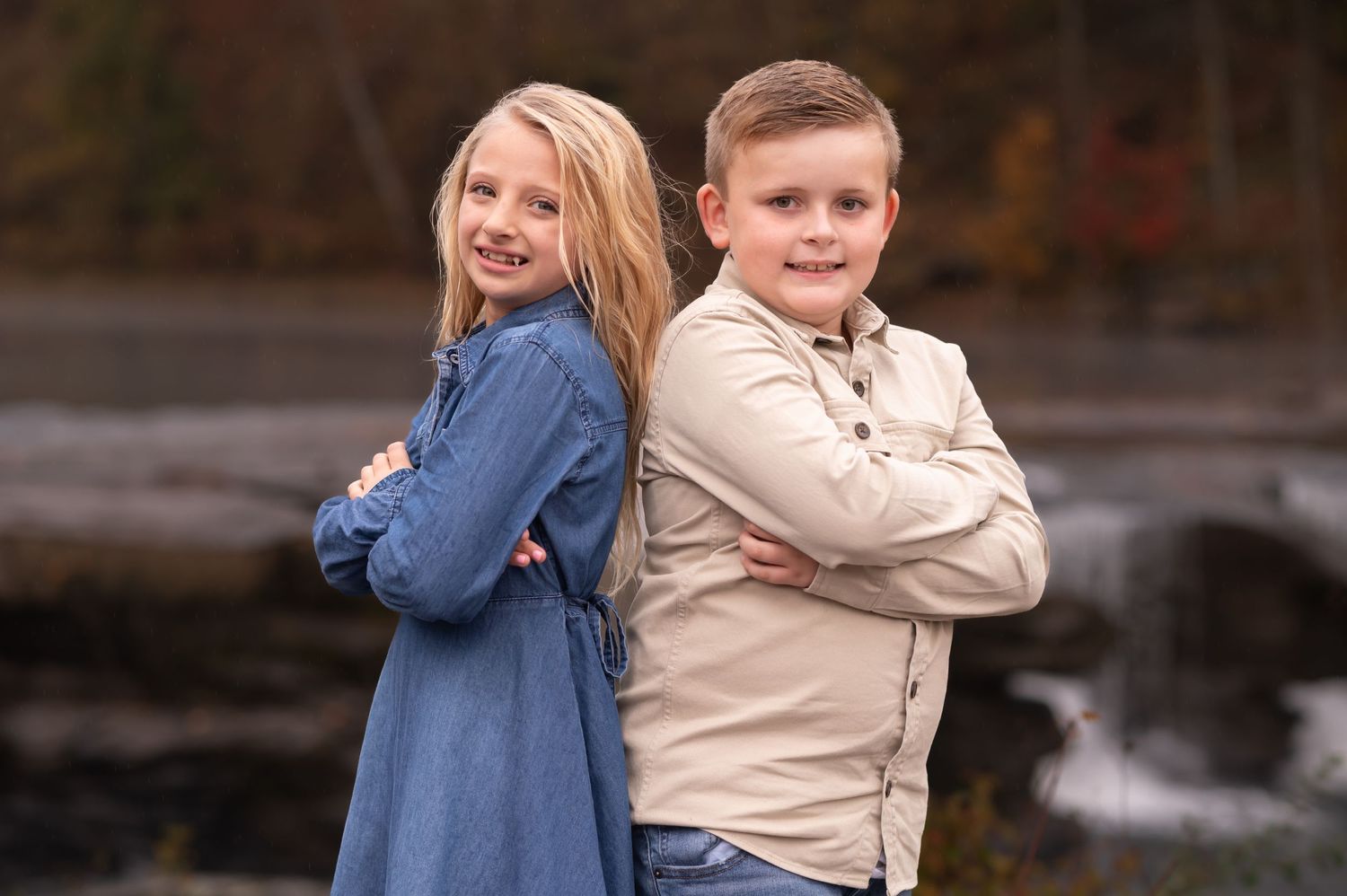 two small siblings pose back to back in front of a waterfall for a portrait. Fall leaves fill the background.