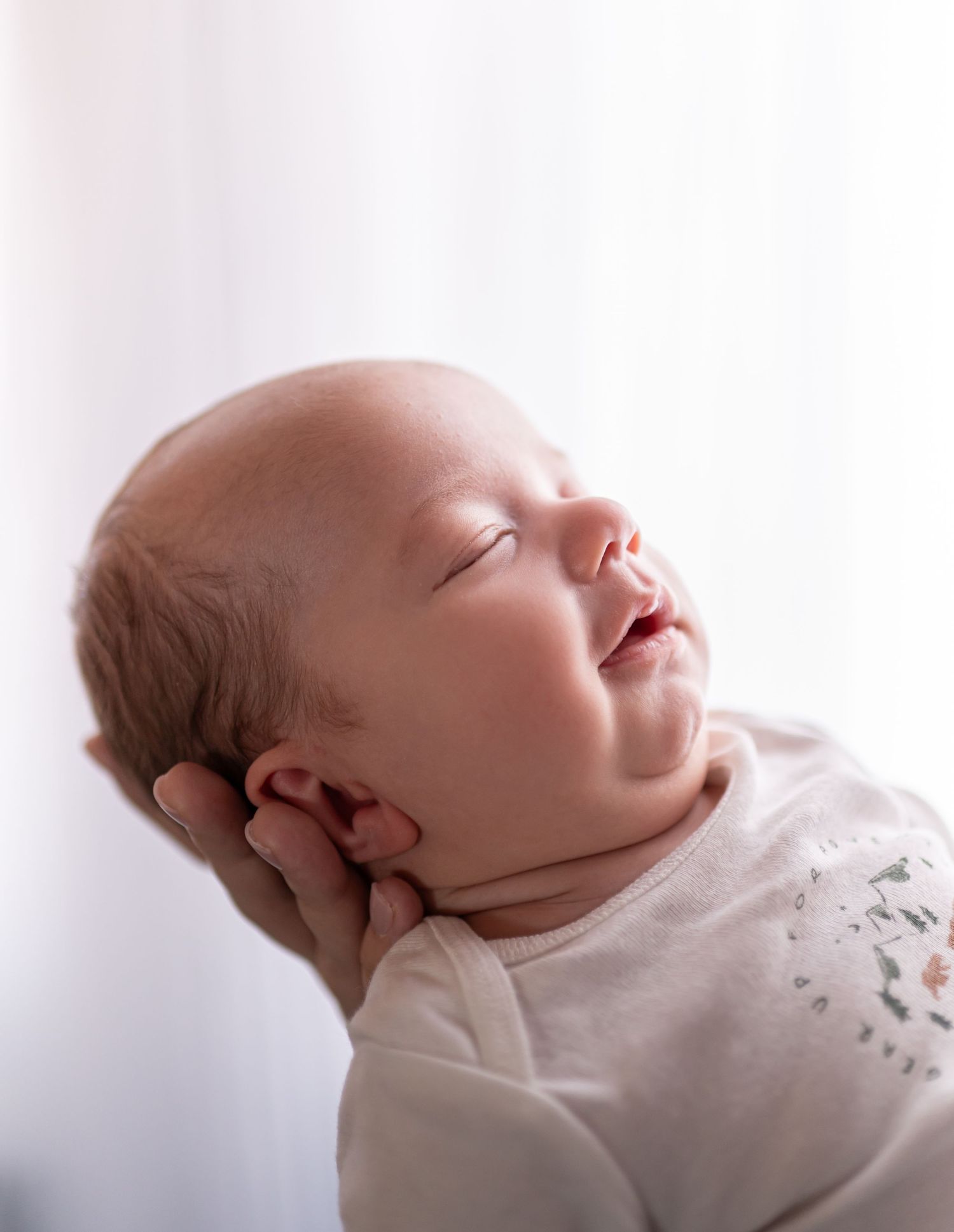 Sleeping newborn baby cradled in a parent's hand, wearing a white onesie, with a peaceful smile on their face.