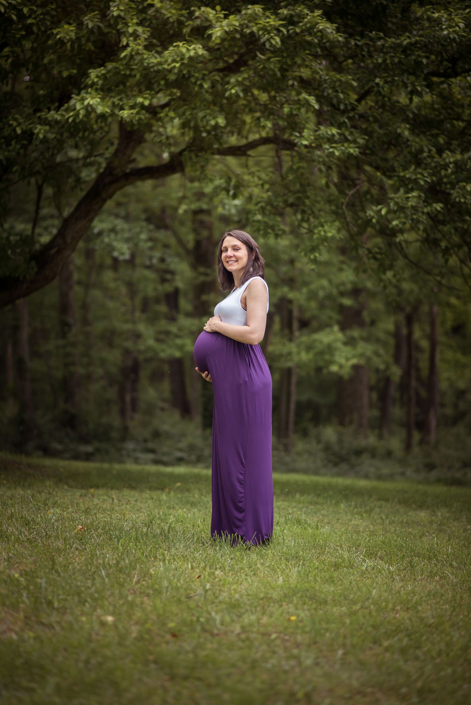 a pregnant woman in a purple and white dress stands under hanging branches in the clearing of a field, with her hands on her belly