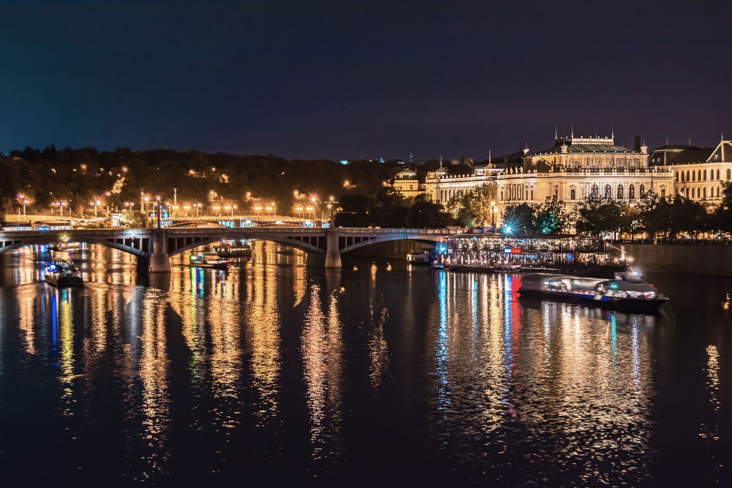 Illuminated bridge and historic buildings reflect on the Vltava River at night in Prague, Czech Republic.