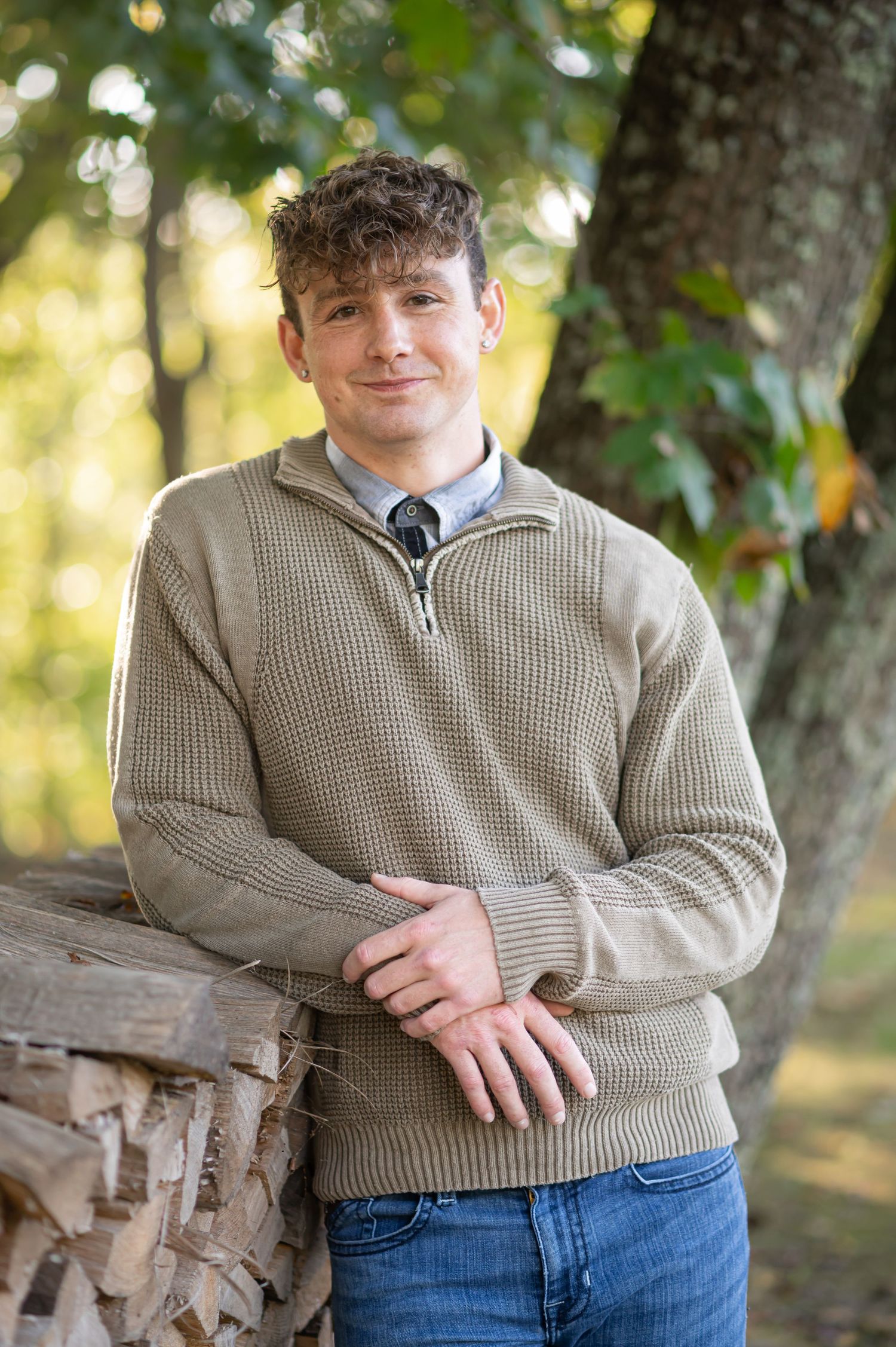 headshot of a young man in a shirt, tie, and brown sweater, leaning against a woodpile and smiling for the camera
