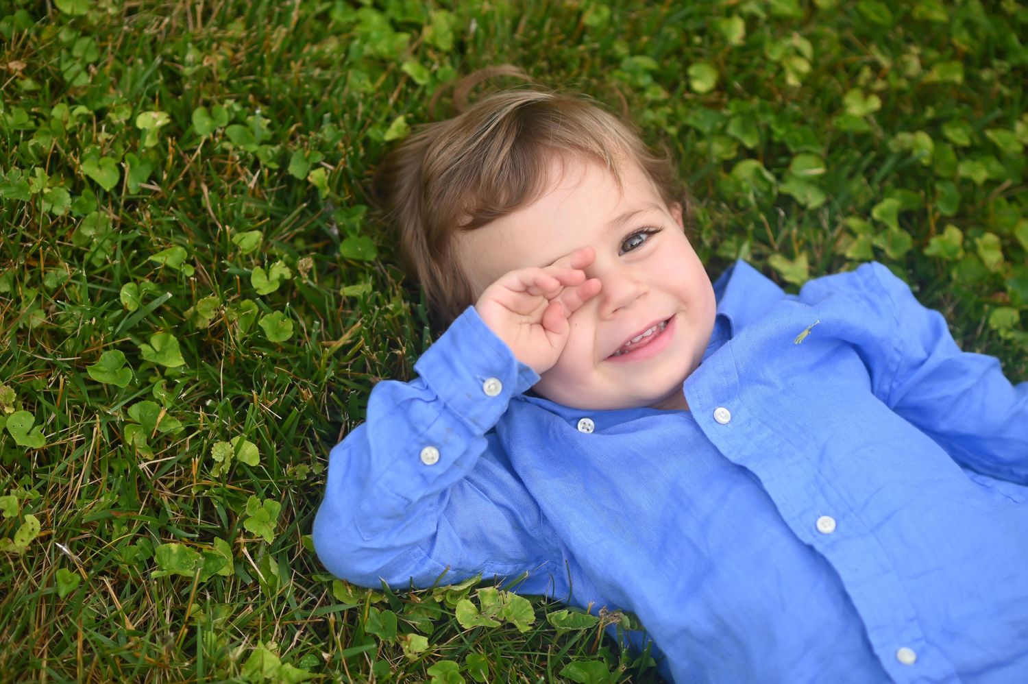 a little boy in a blue shirt lies in the grass and laughs as he peeks out from behind one hand at the camera