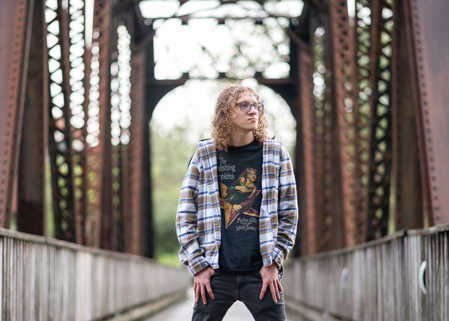 a young man with long curly hair, in a band tee and plaid shirt, poses with a wide stance on an old iron bridge in downtown Morgantown WV