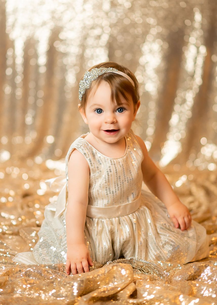 a toddler girl with brown hair in a sparkly gold sequin dress and a shimmering headband kneels and smiles on a backdrop of gold sequins