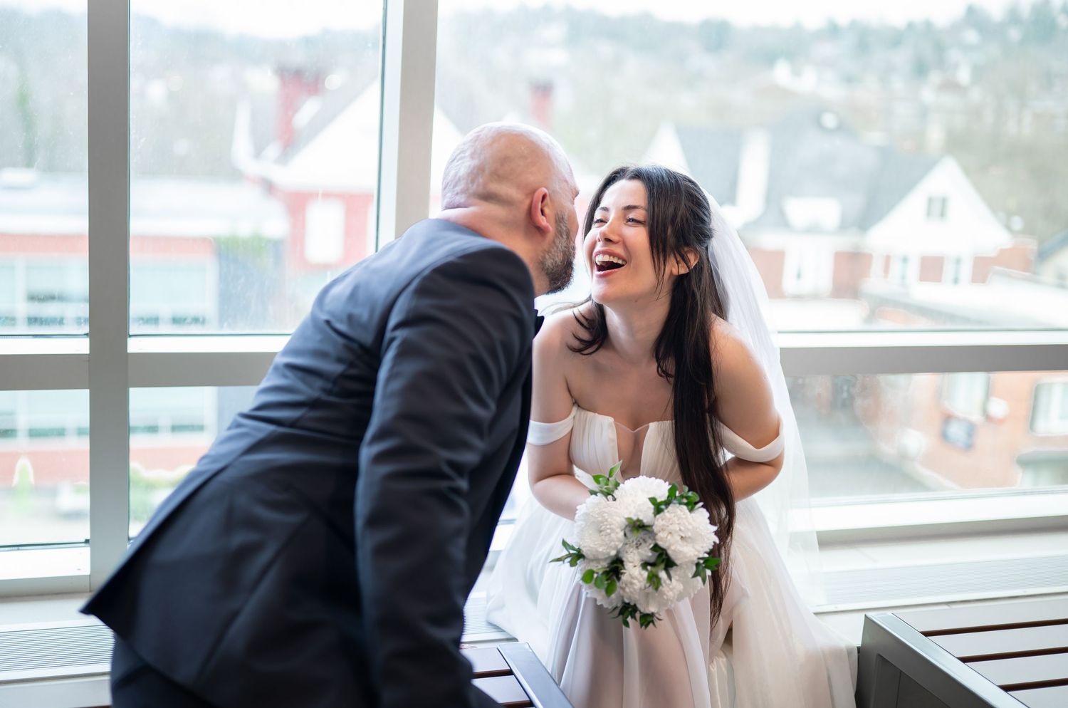 Candid portrait of a bride and group sitting in front of large windows overlooking downtown Morgantown, talking and laughing