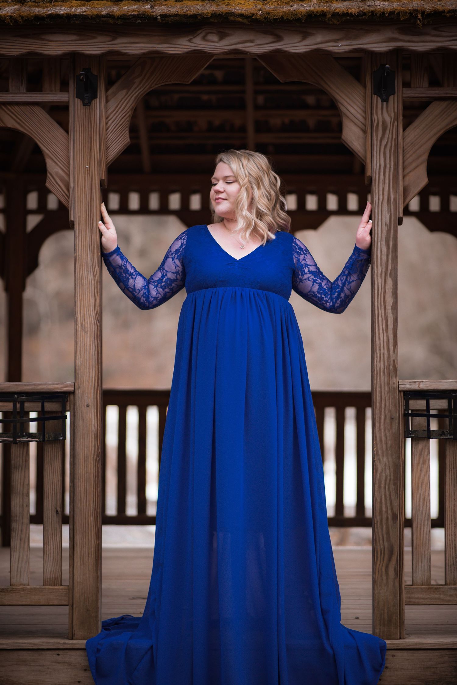 a pretty young blonde pregnant woman poses in a   wood gazebo, in a dramatic blue gown with waterfalls behind her.