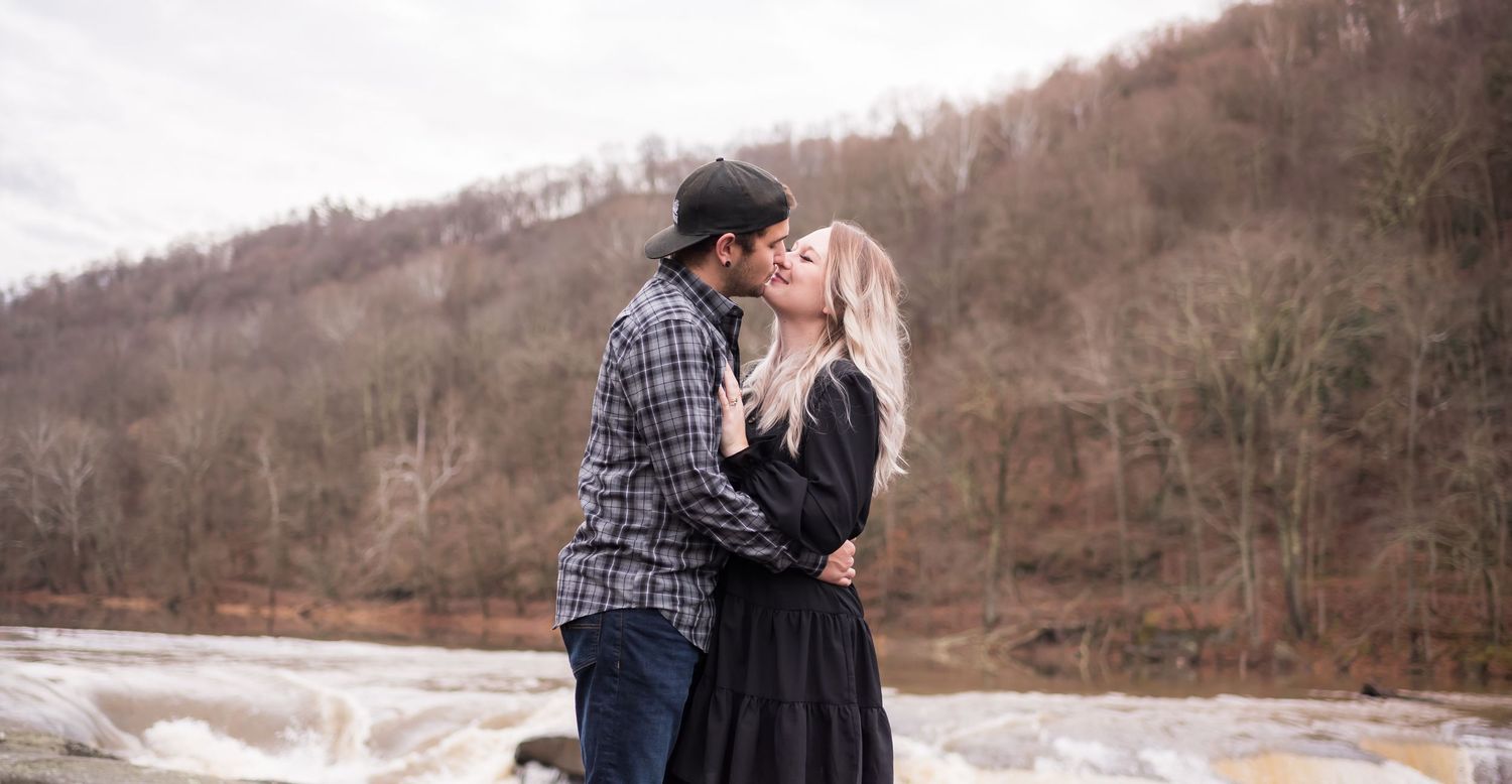 a young couple embraces in front of a roaring river with fall scenery in the background, at Valley Falls State Park in Morgantown, WV