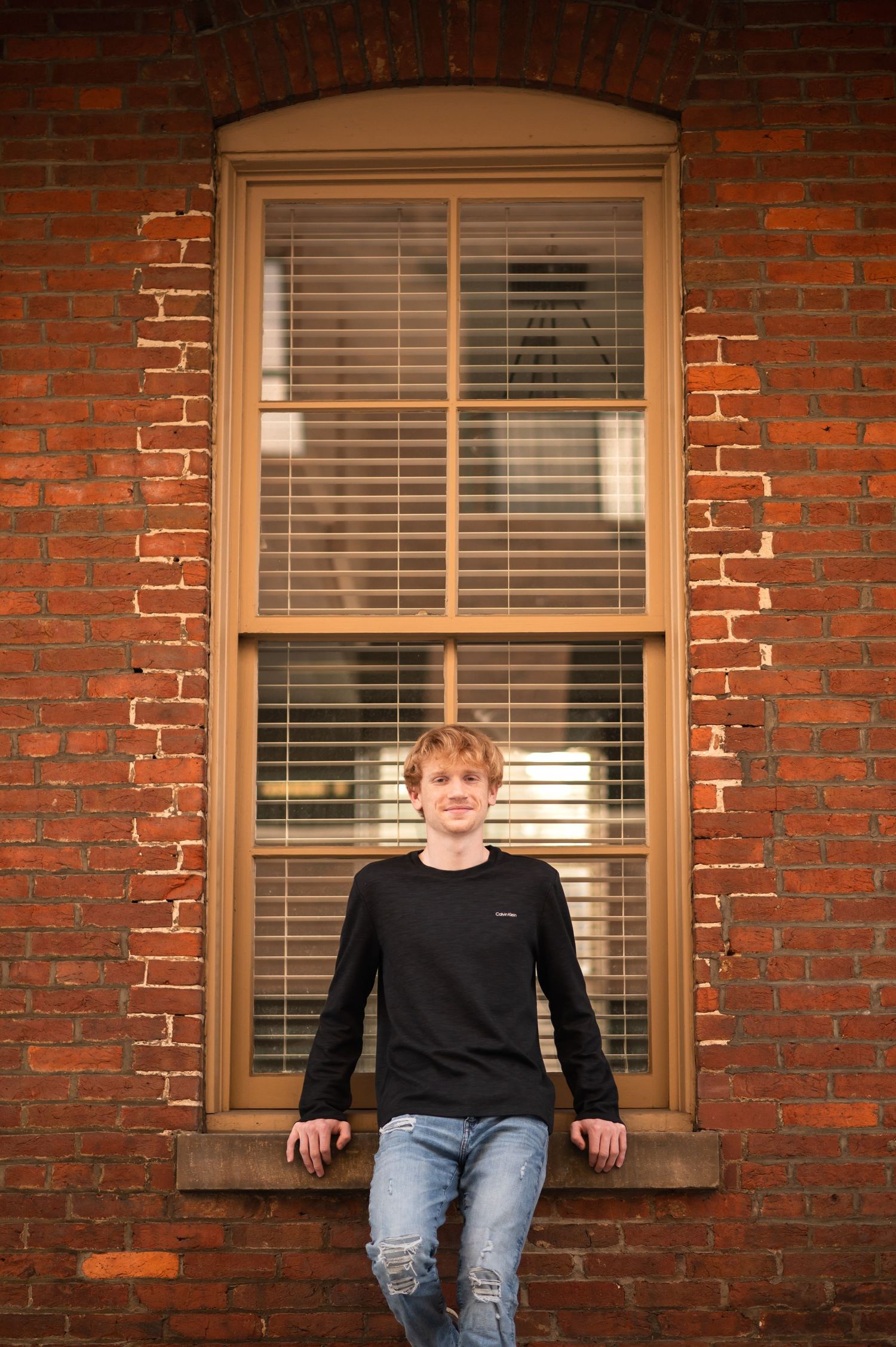 a senior boy in a black shirt and jeans lounges against a tall old window in a brick wall, in downtown Morgantown, WV