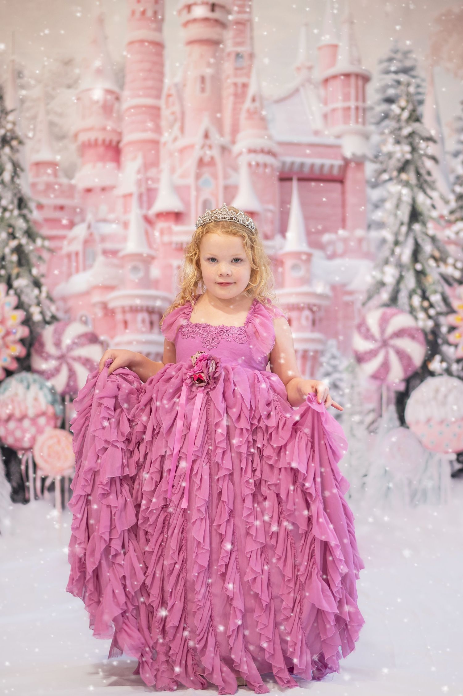 a little girl in a frilly pink dress poses in front of a pink candy christmas castle scene in the snow