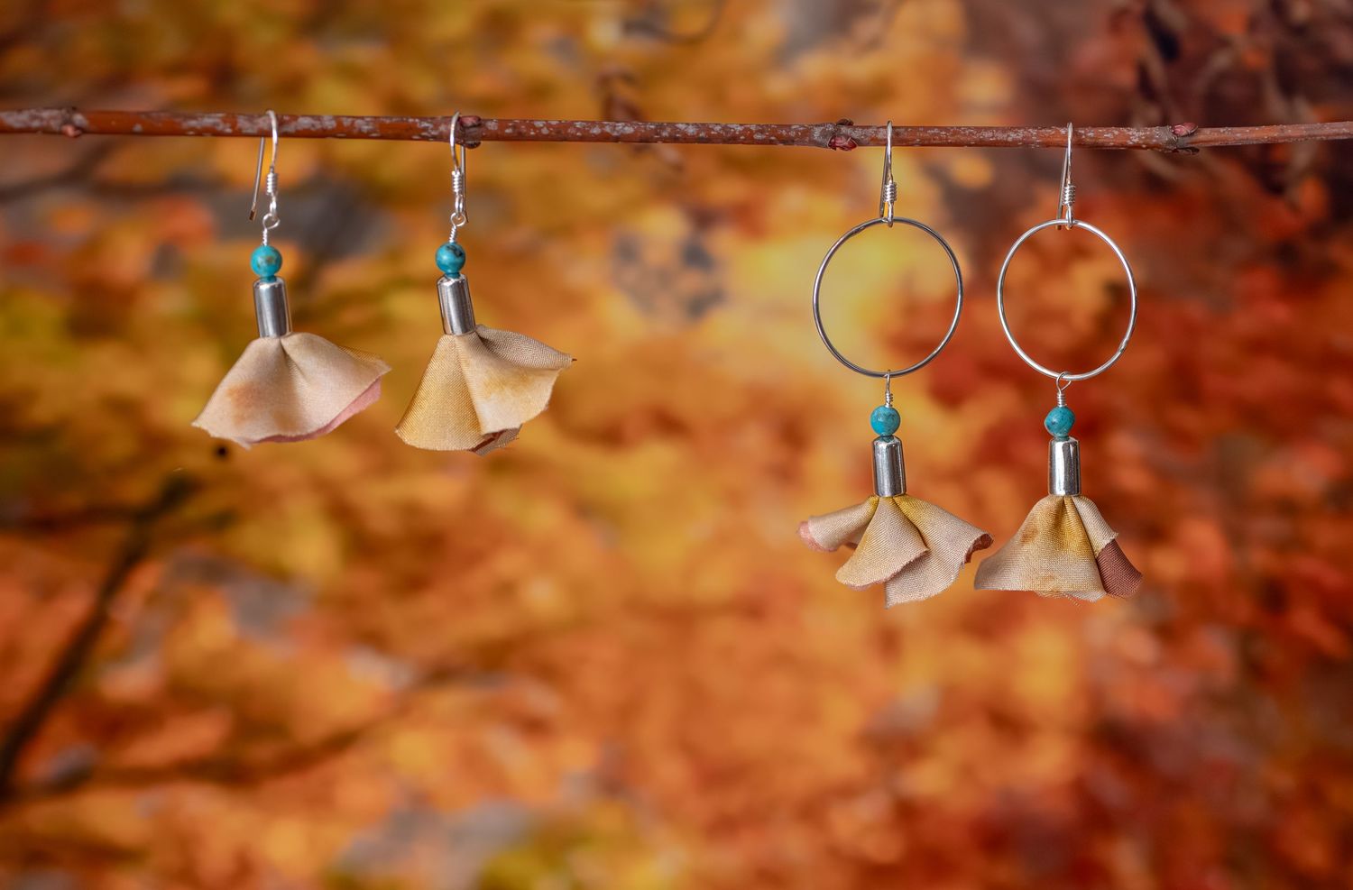 Two pairs of handmade brass and turquoise dangle earrings hanging on a branch with autumn foliage backdrop during a commercial shoot