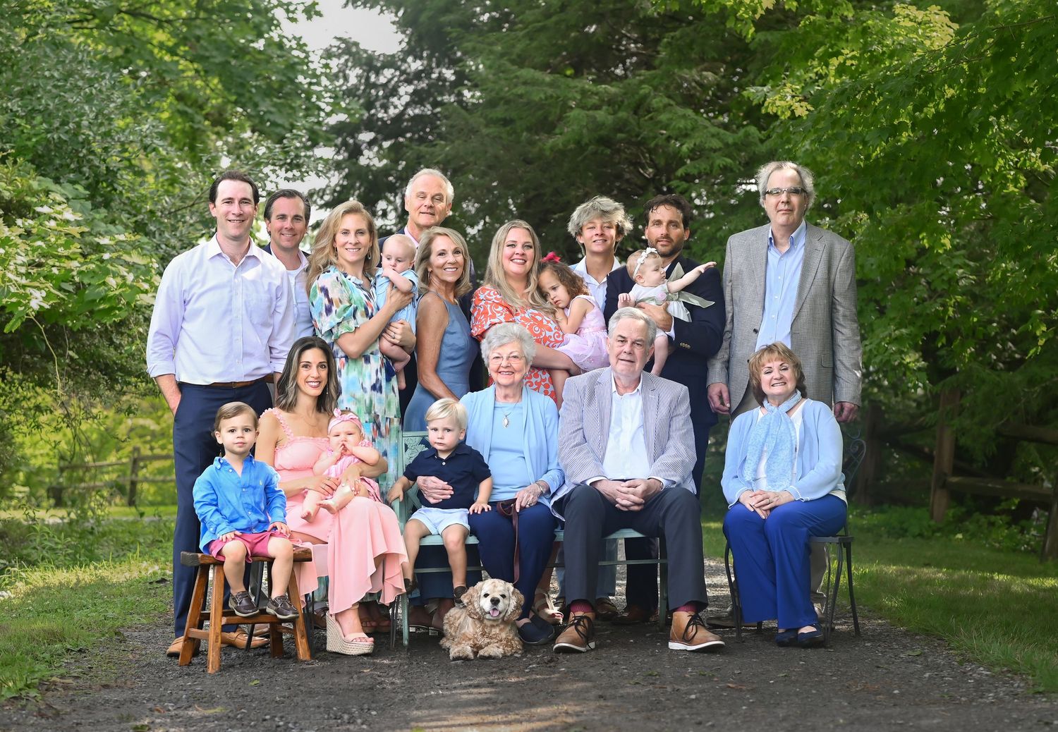 an extended family poses together on an old country road with trees and grass in the background for a four-generations picture