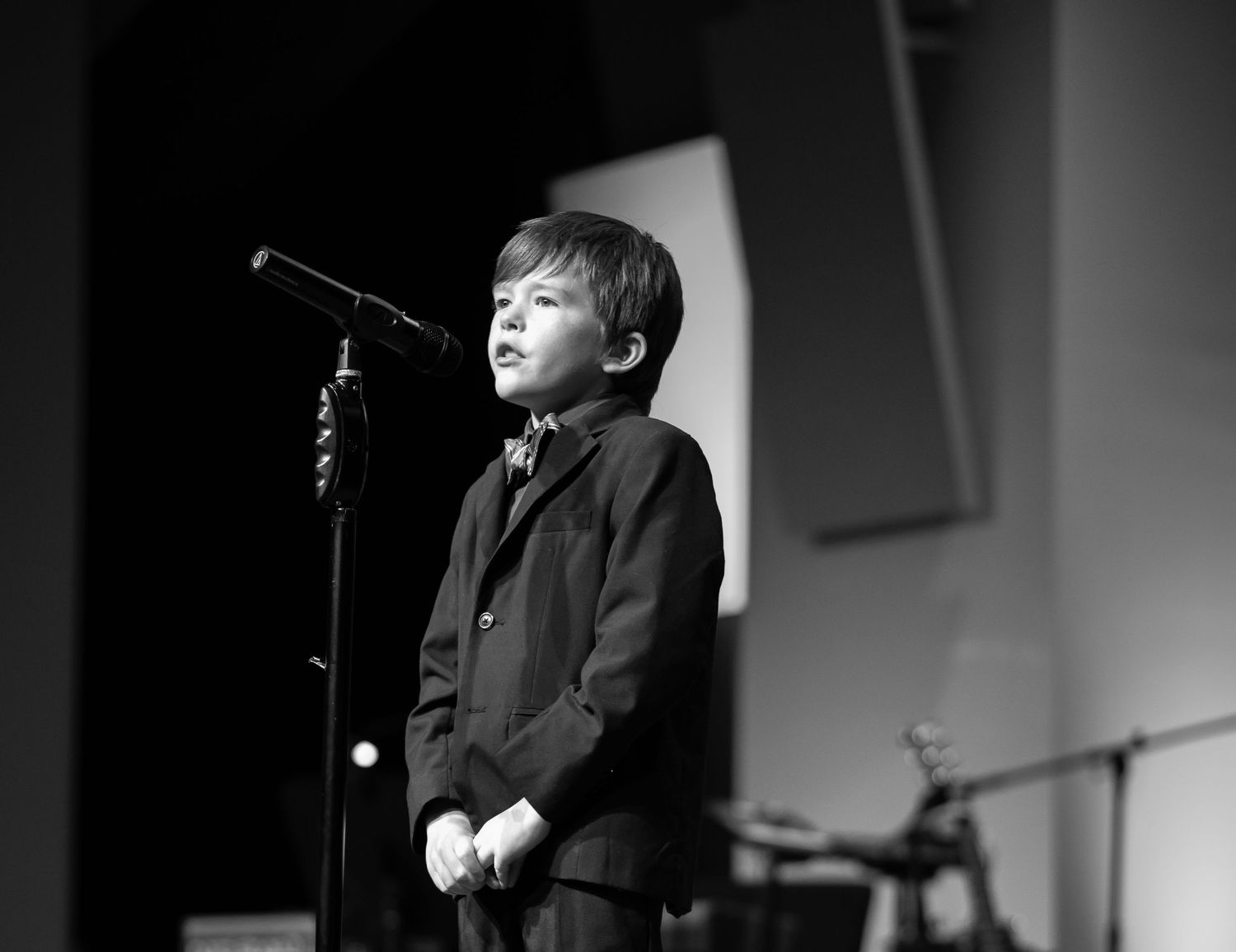 candid black and white image of a boy in a suit and tie, standing nervously at a microphone in a large auditorium