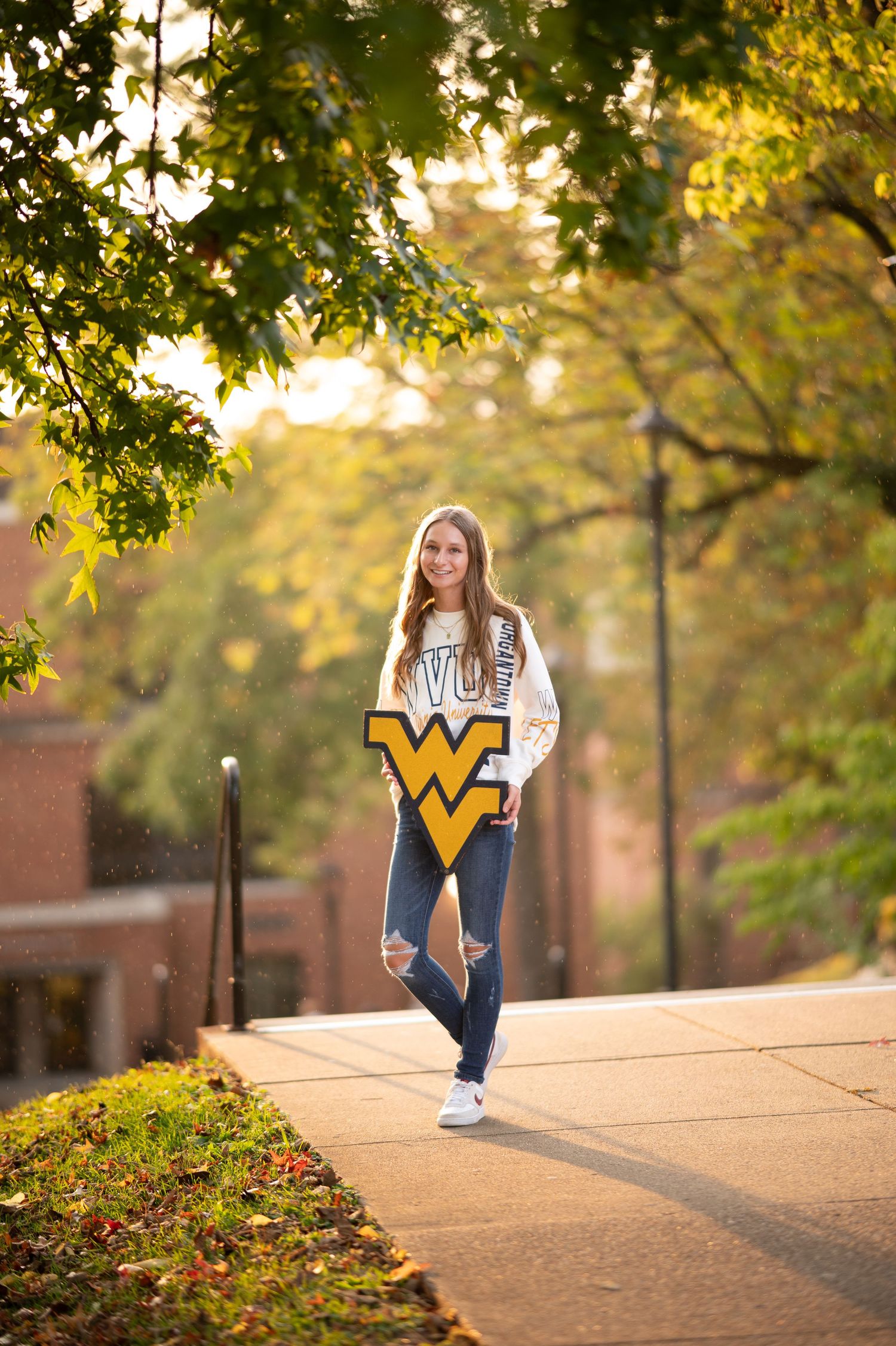 a senior student in a WVU sweatshirt holds a blue and gold WVU sign and stands in the sun on WVU's campus with rain falling around her