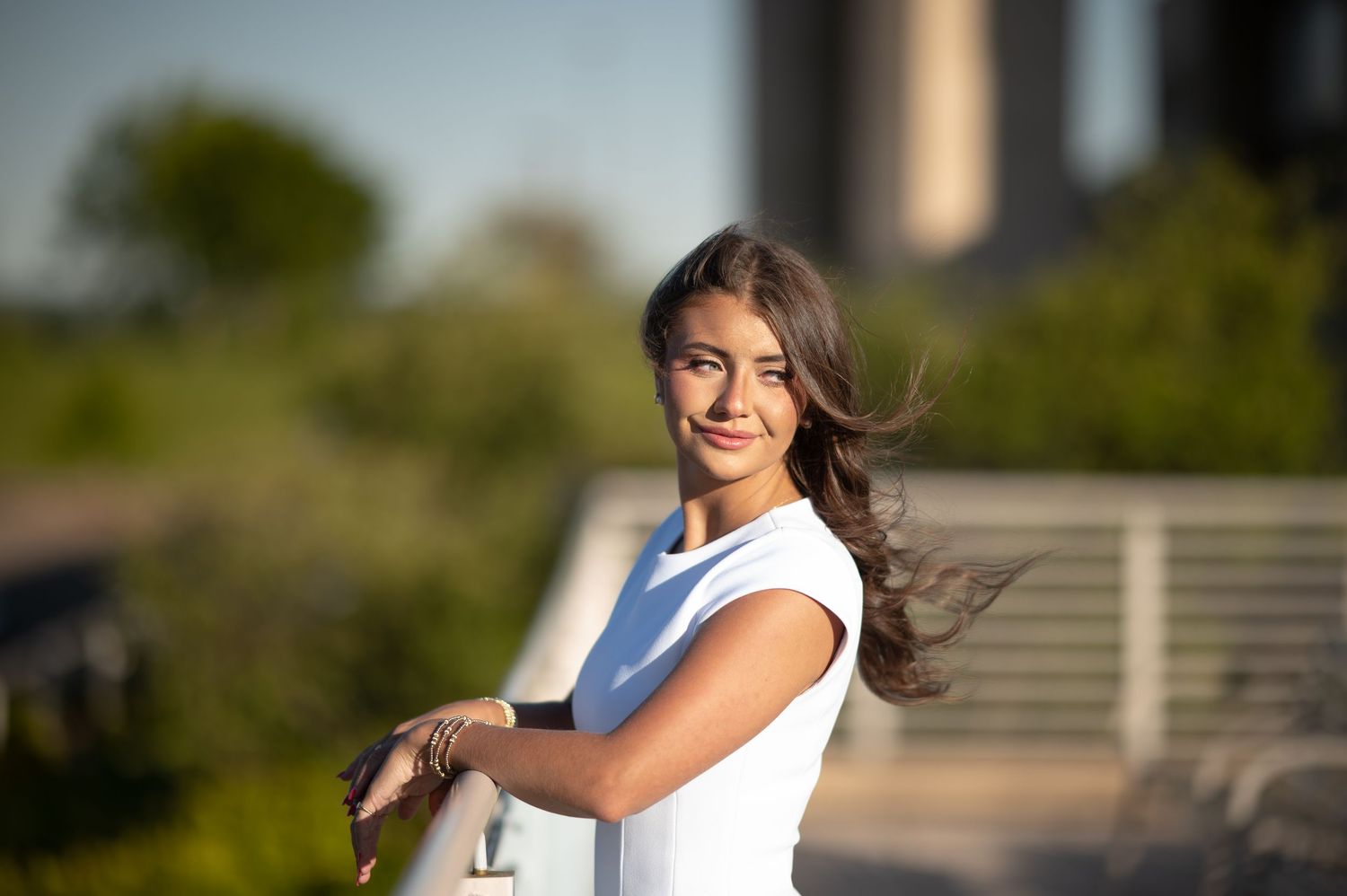 a young woman in a white dress leans against a railing and smiles as the wind blows her hair
