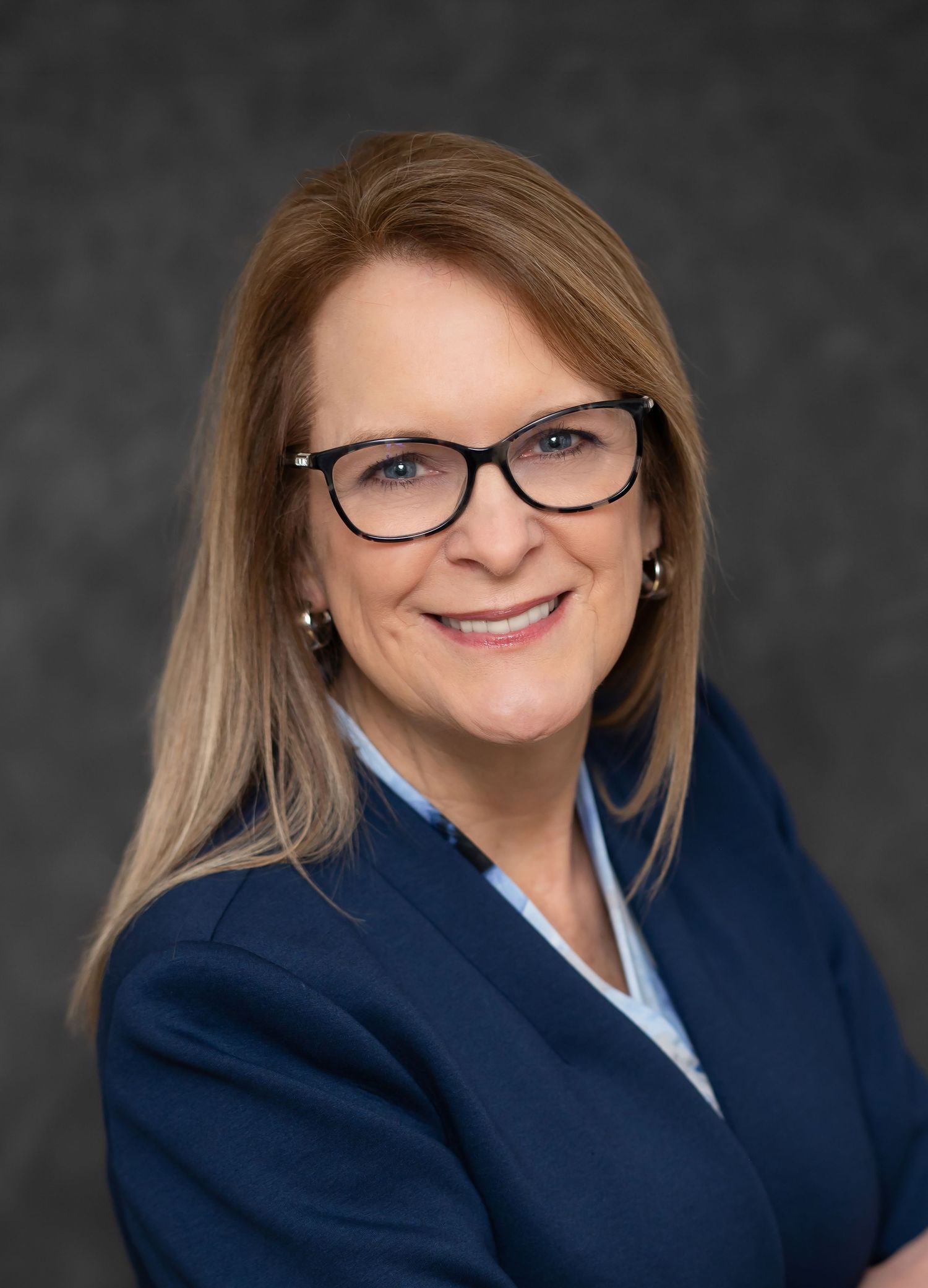 a woman with light brown hair and glasses in a navy suit smiles into the camera for a headshot against a dark background