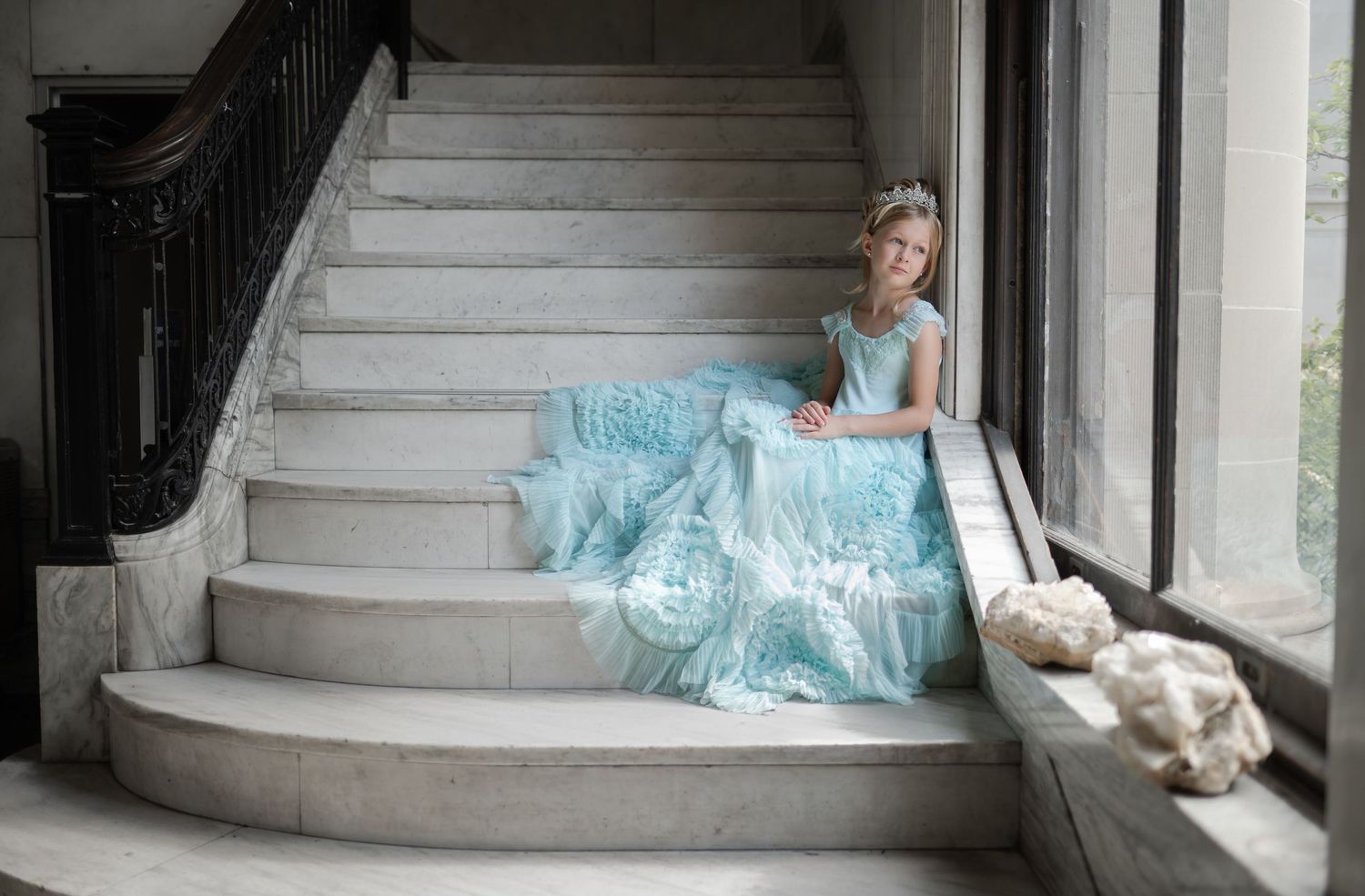 Portrait of a young girl in a light blue ball gown sitting gracefully on marble stairs near a window.
