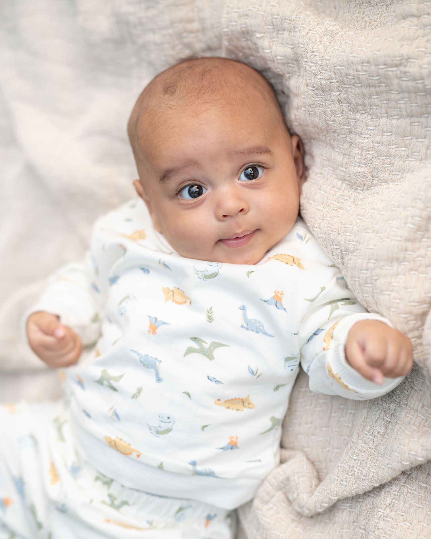 a little boy with big dark brown eyes in a white onesie lies on a tan blanket and looks into the camera