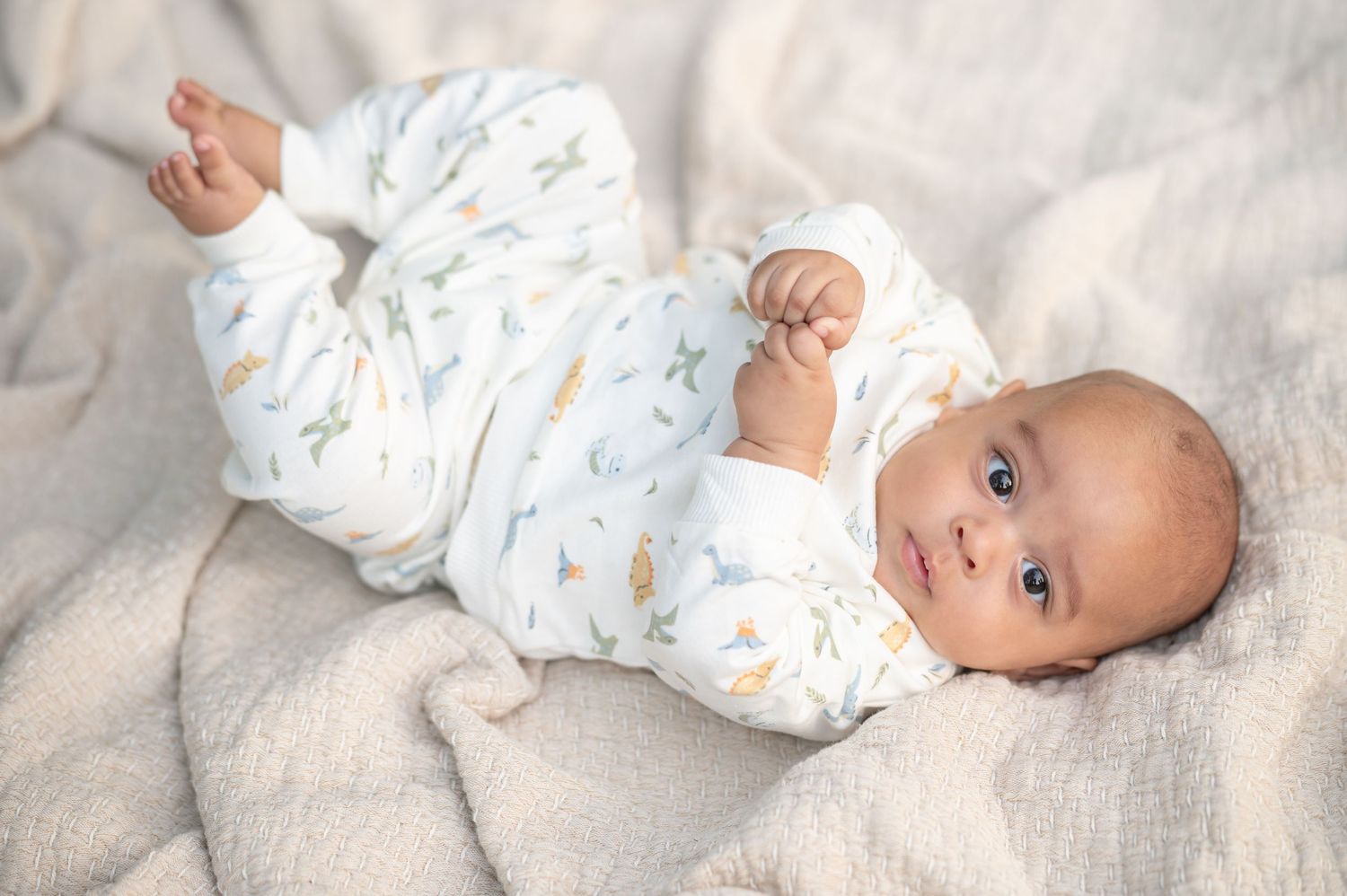a baby boy with big dark eyes in a white romper lies on a tan blanket at looks into the camera