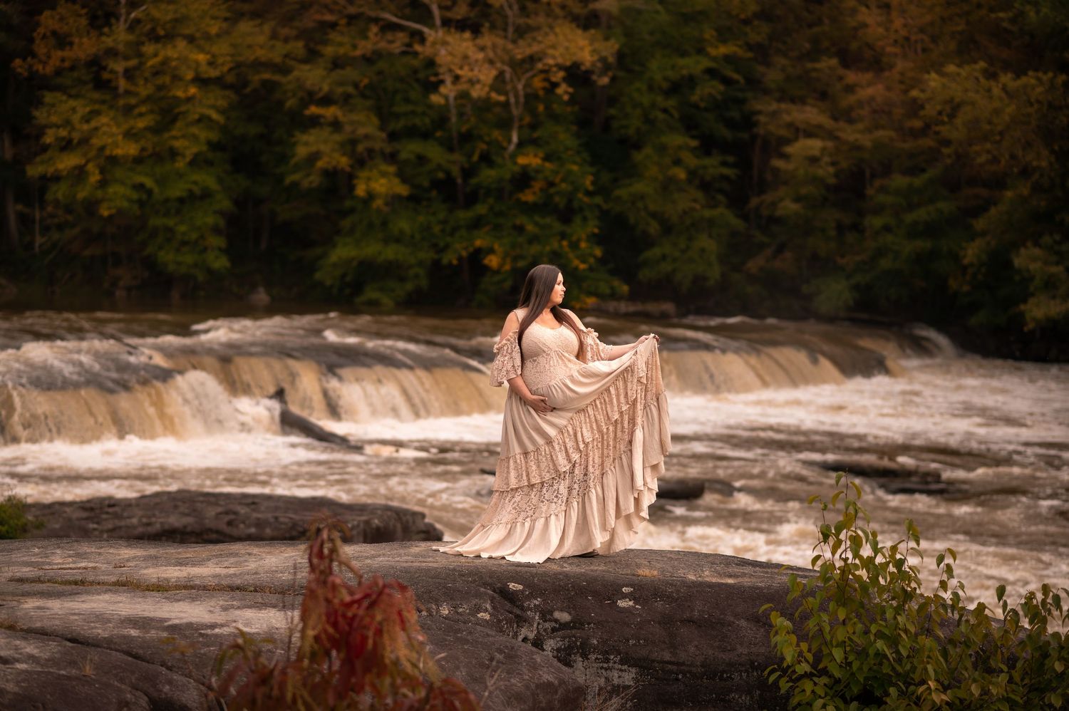 a pregnant woman in a long beige lace dress stands on a large rock in front of the roaring waterfalls at Valley Falls State Park, in WV