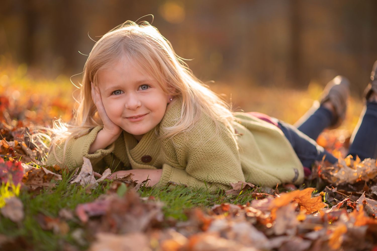 a little blonde girl in a green sweater lies in the grass surrounded by brilliant fall leaves and smiles for a photo