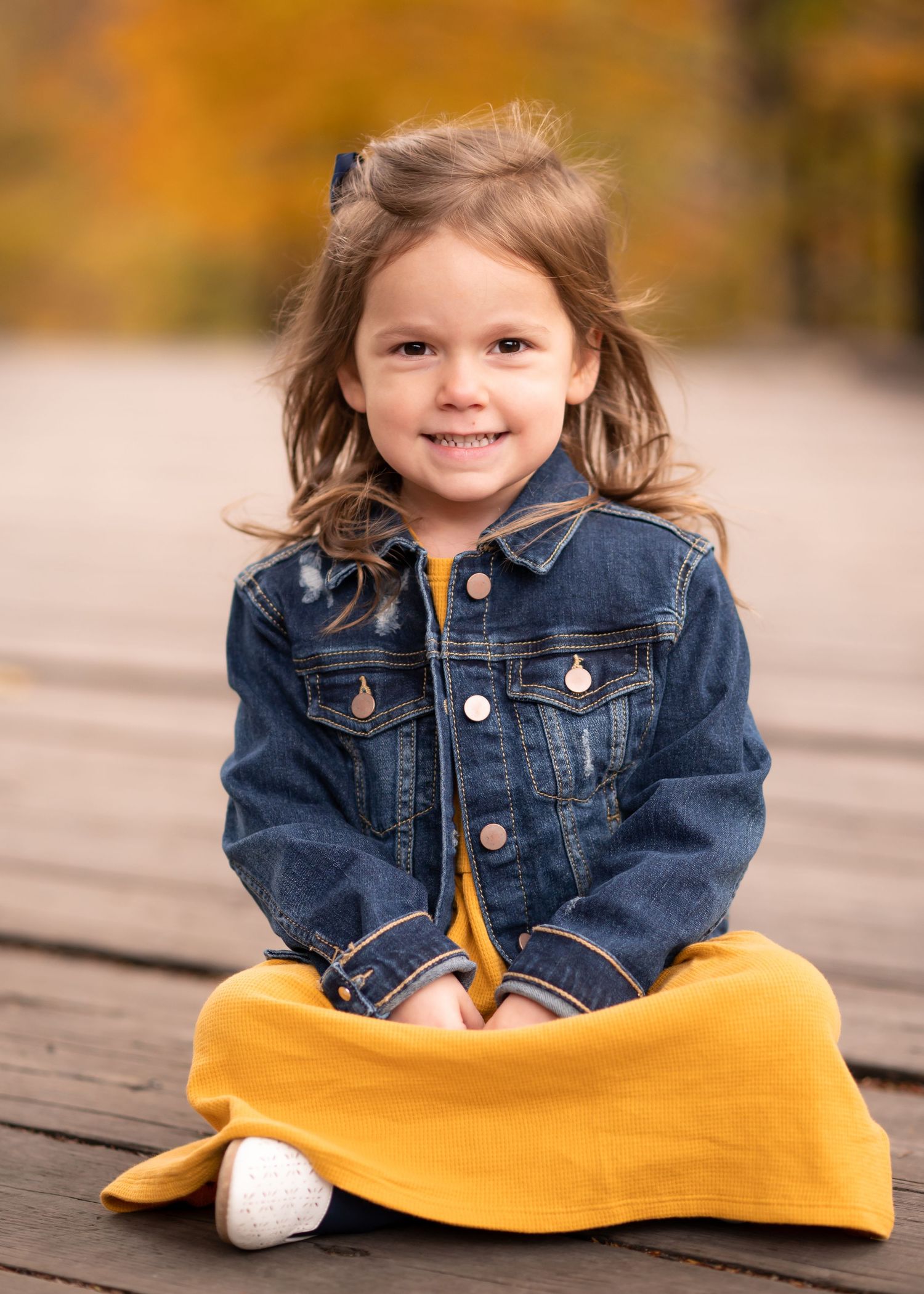 a little girl in a yellow dress and a jean jacket sits crosslegged on an old wooden bridge and smiles at the camera for a portrait