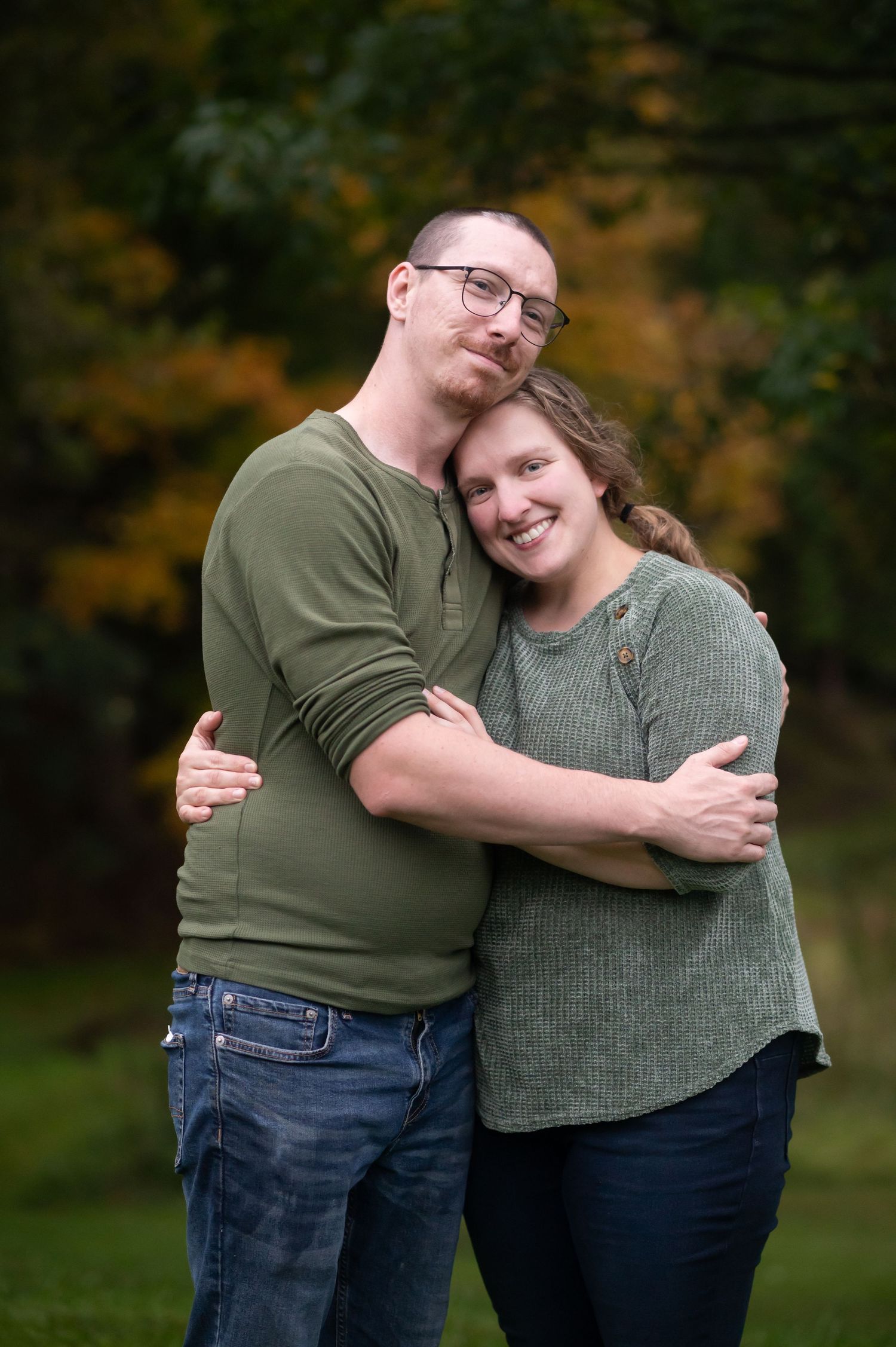 a husband and wife embrace and smile for an outdoor portrait with fall scenery in the background