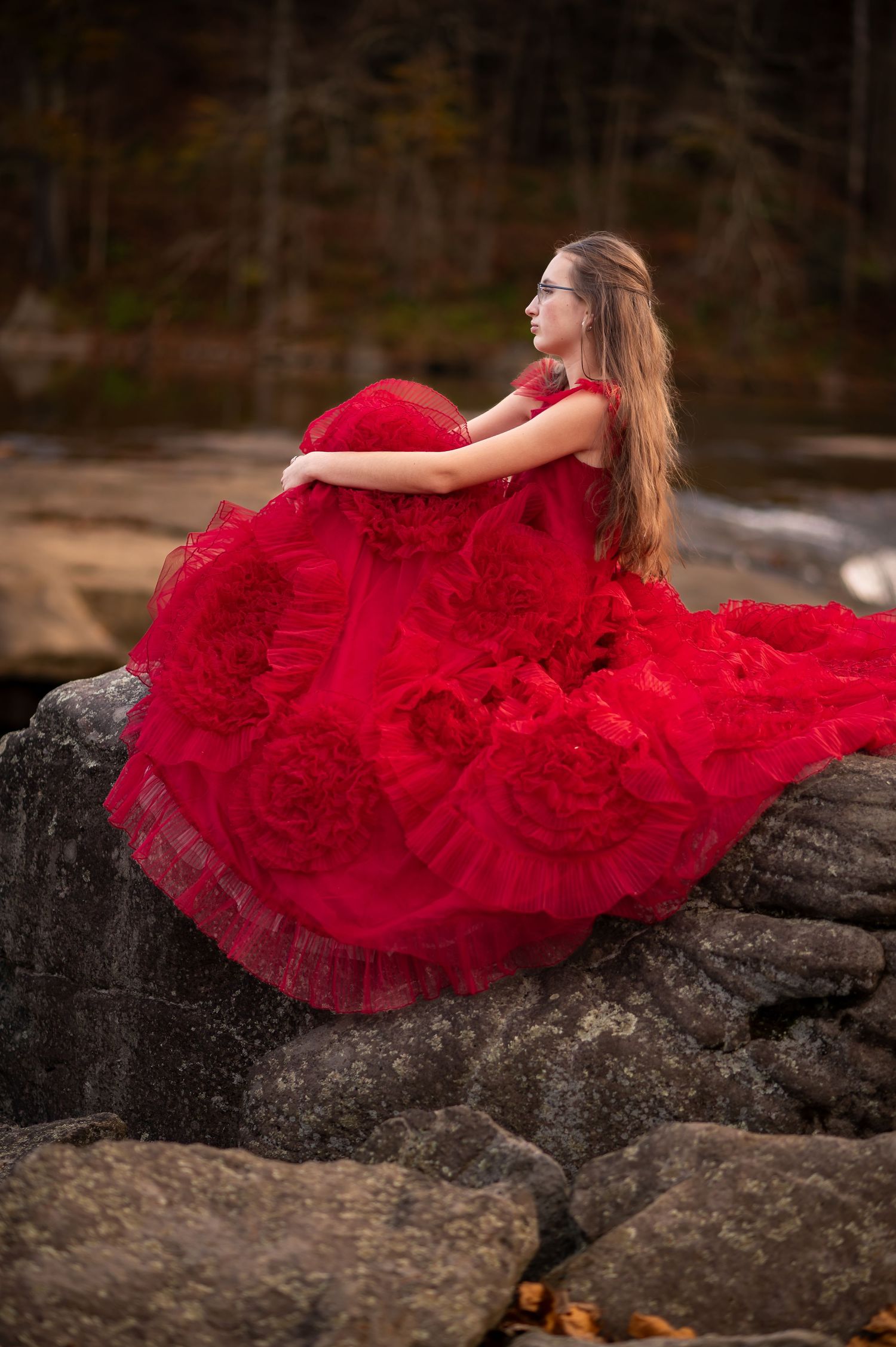 a young woman in a dramatic red dress poses on large rocks in front of a waterfall with the Tygart River behind her at Valley Falls, WV